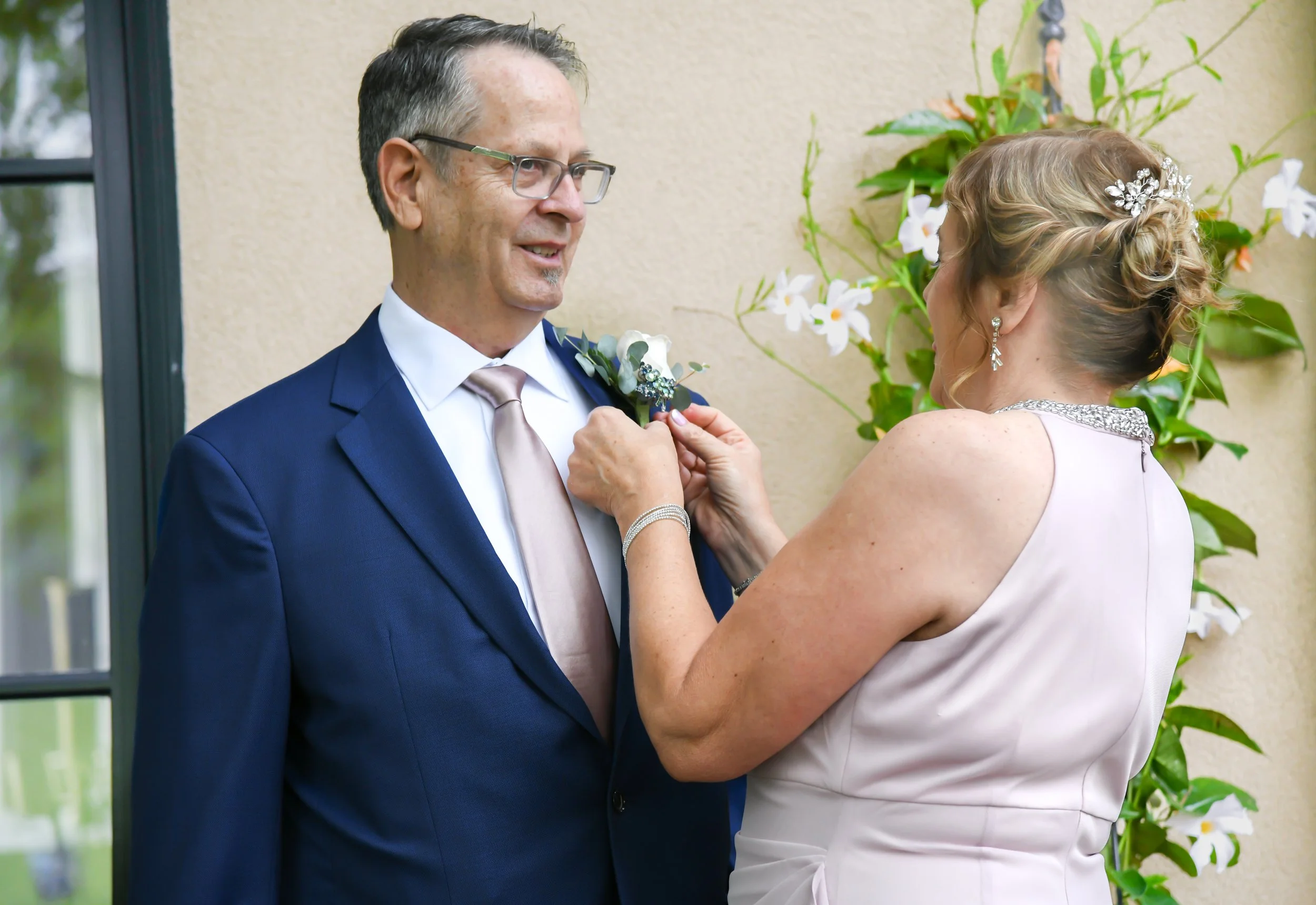 A woman in a light pink dress pins a boutonniere on a man in a navy blue suit at a wedding ceremony.