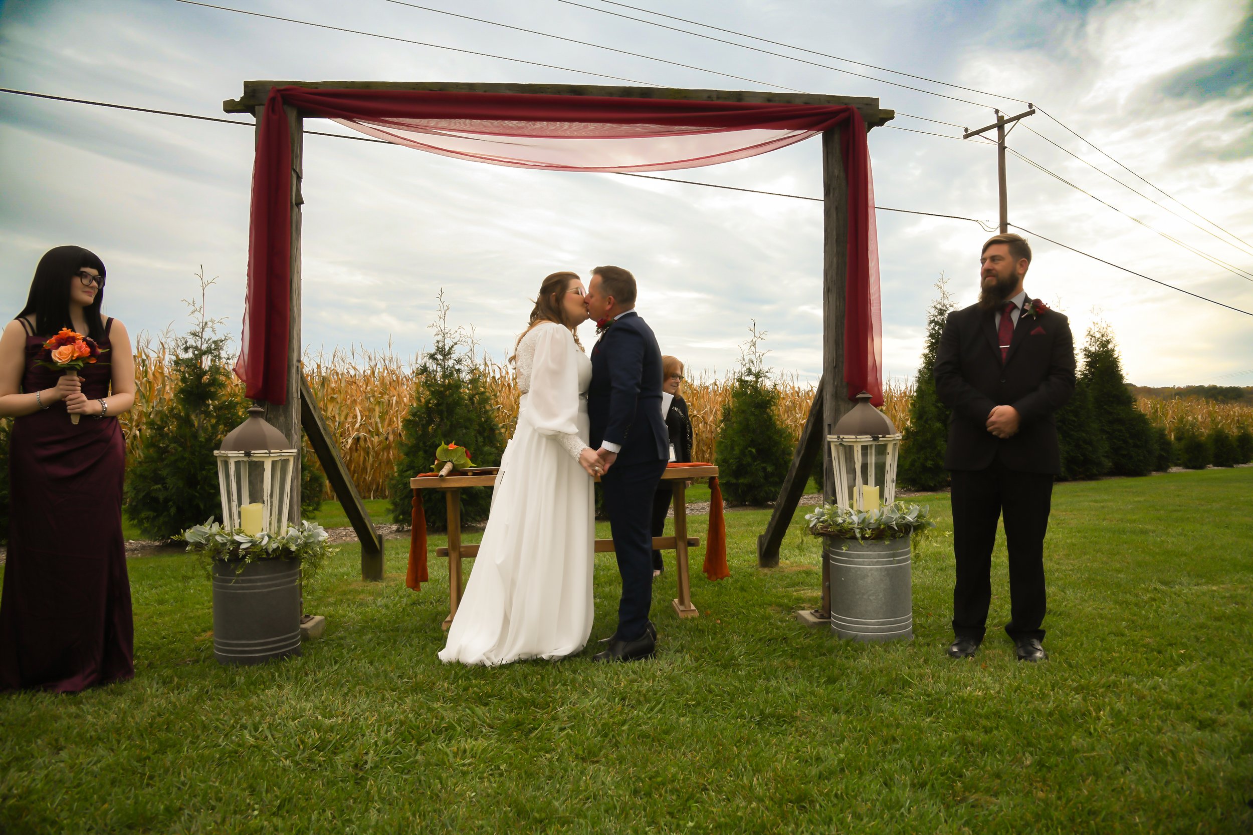 A couple getting married outdoors, kissing under an arch decorated with fabric. The bride is wearing a white dress, and the groom is in a navy suit. Two women and a man stand nearby, with two large lanterns on the grass.
