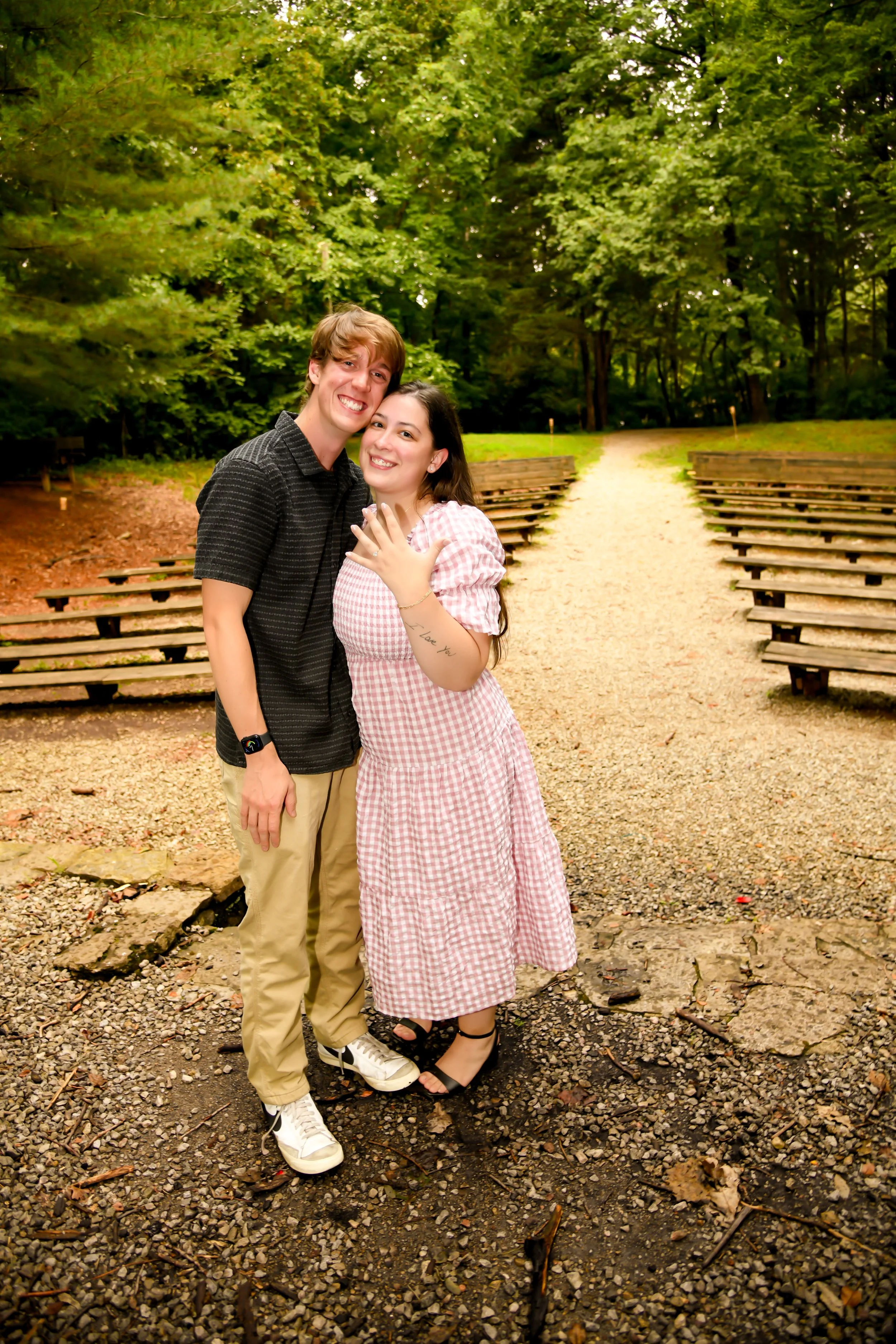 A smiling young man and woman hugging each other in a park with lush green trees and benches in the background, on a gravel pathway.