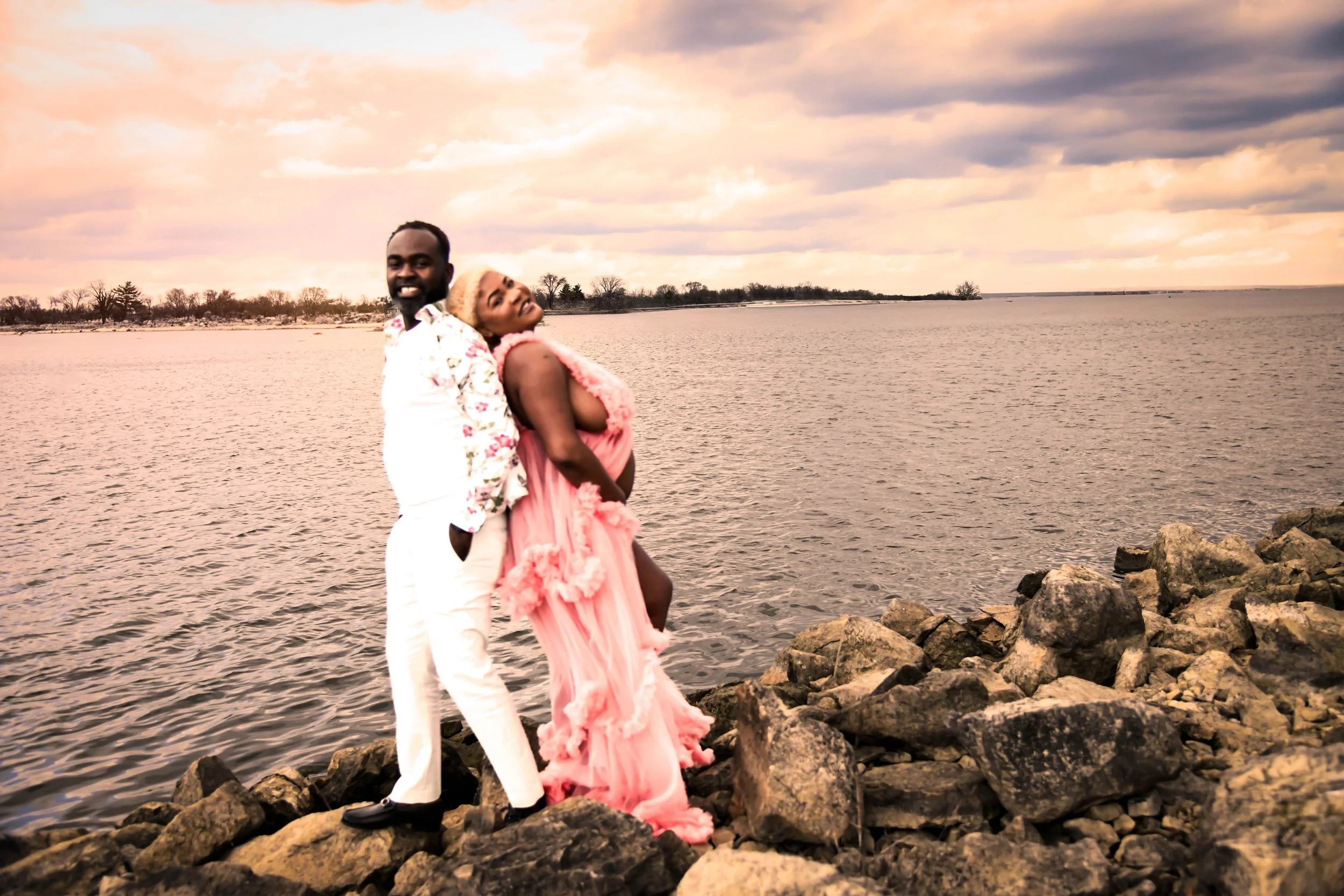 A couple standing on rocks by a body of water during sunset, with the man wearing a white suit and the woman in a pink dress.