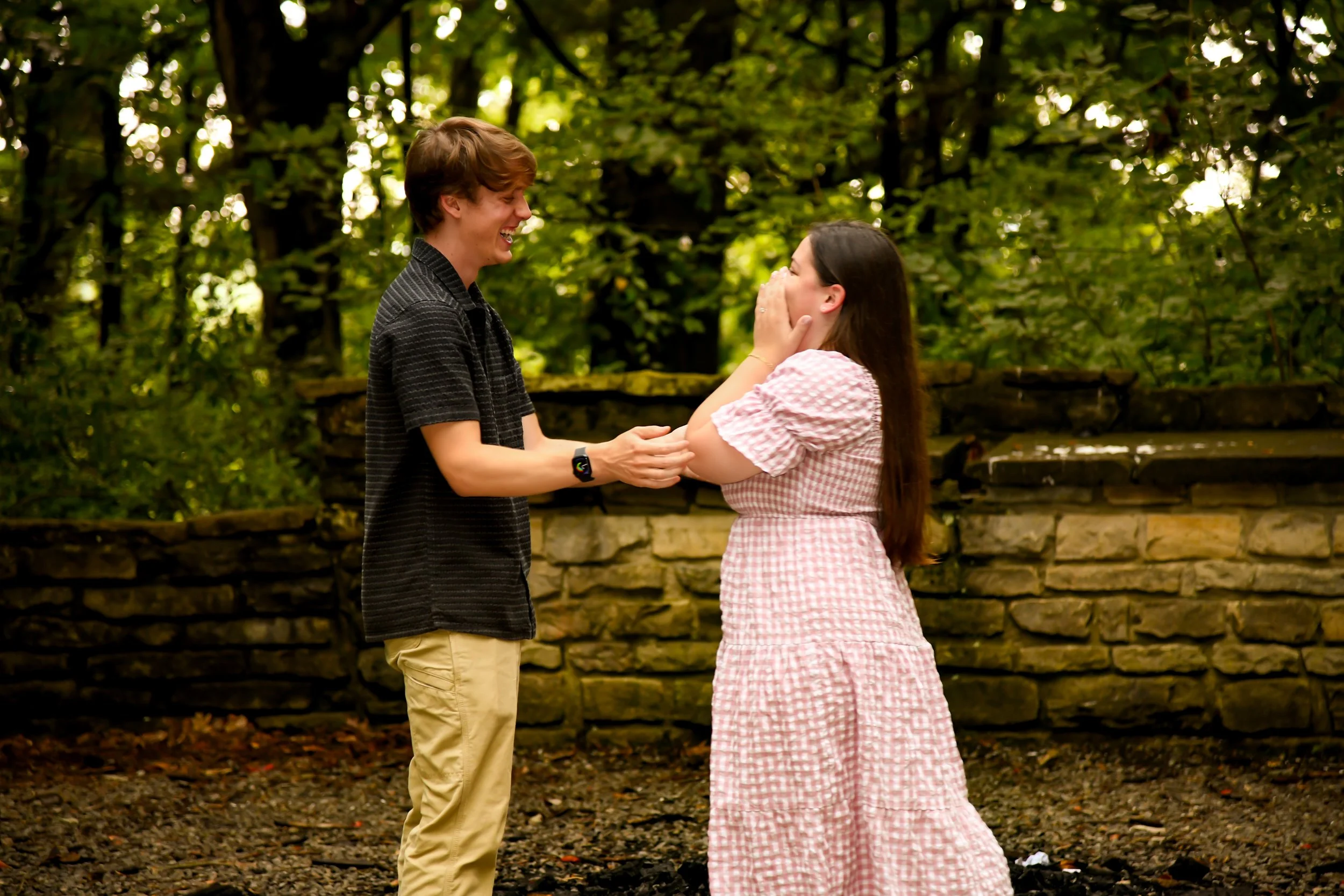 A young man and woman sharing a joyful moment outdoors, with the man holding the woman's arms, both smiling and laughing in front of a stone wall and trees.