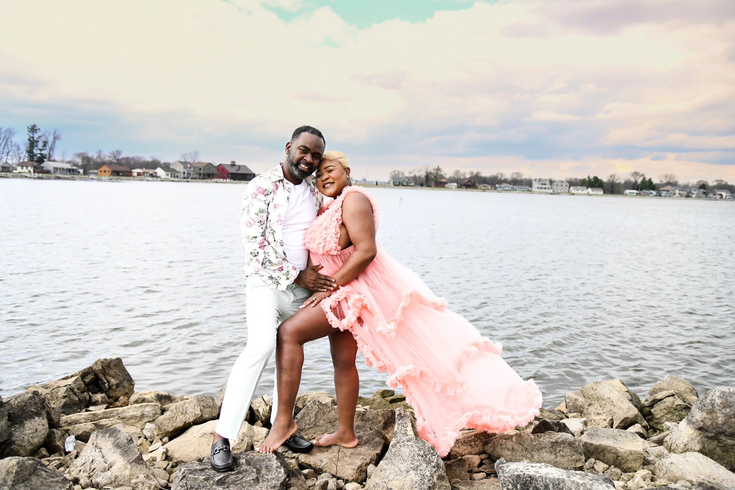 A man and woman standing on rocks by a body of water, embracing and smiling, with a residential neighborhood in the background.