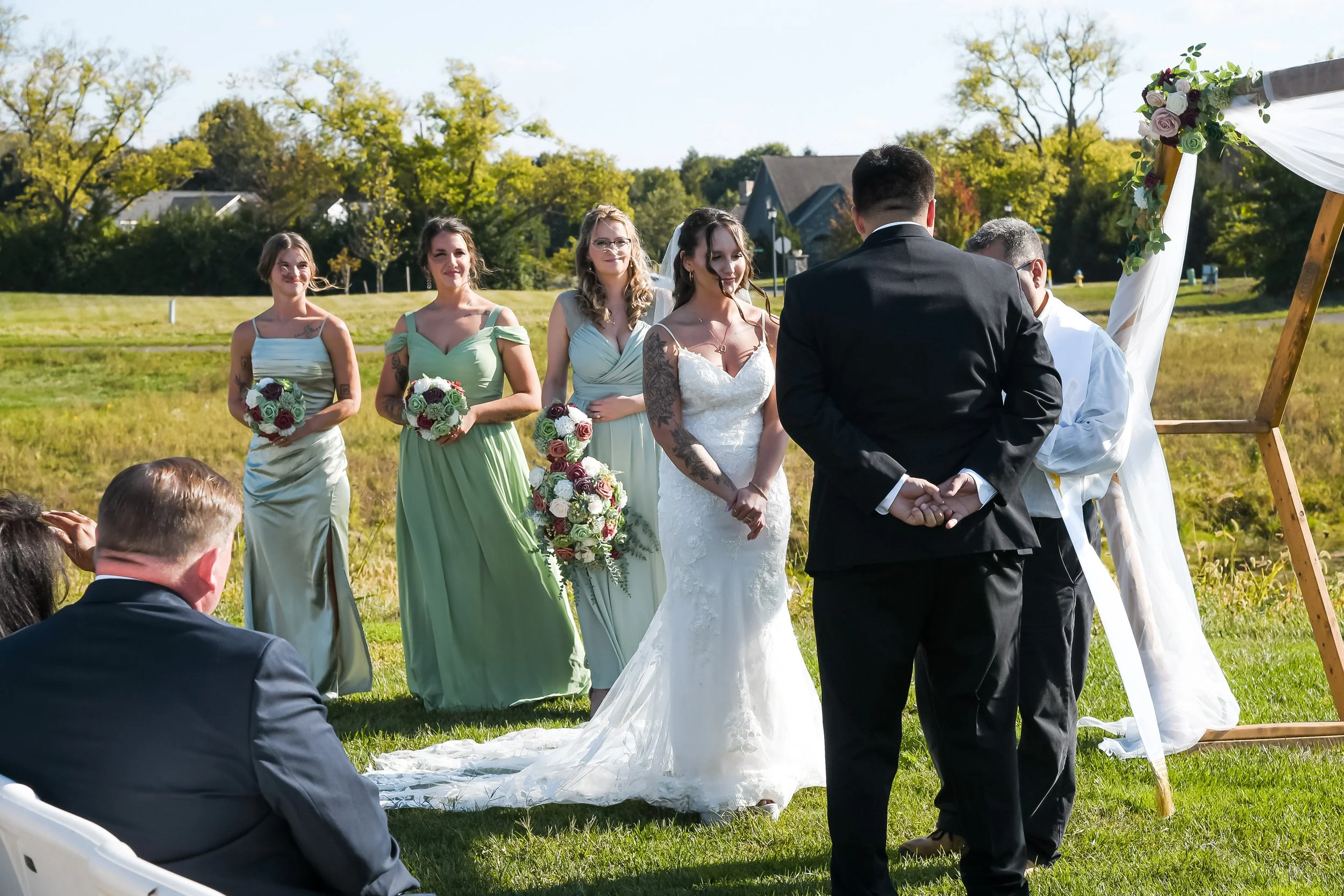 A couple gets married outdoors with five women dressed in pastel-colored bridesmaid dresses standing behind them. The bride and groom stand in front of a wooden wedding arch decorated with flowers and white fabric, on a grassy field with trees and ho