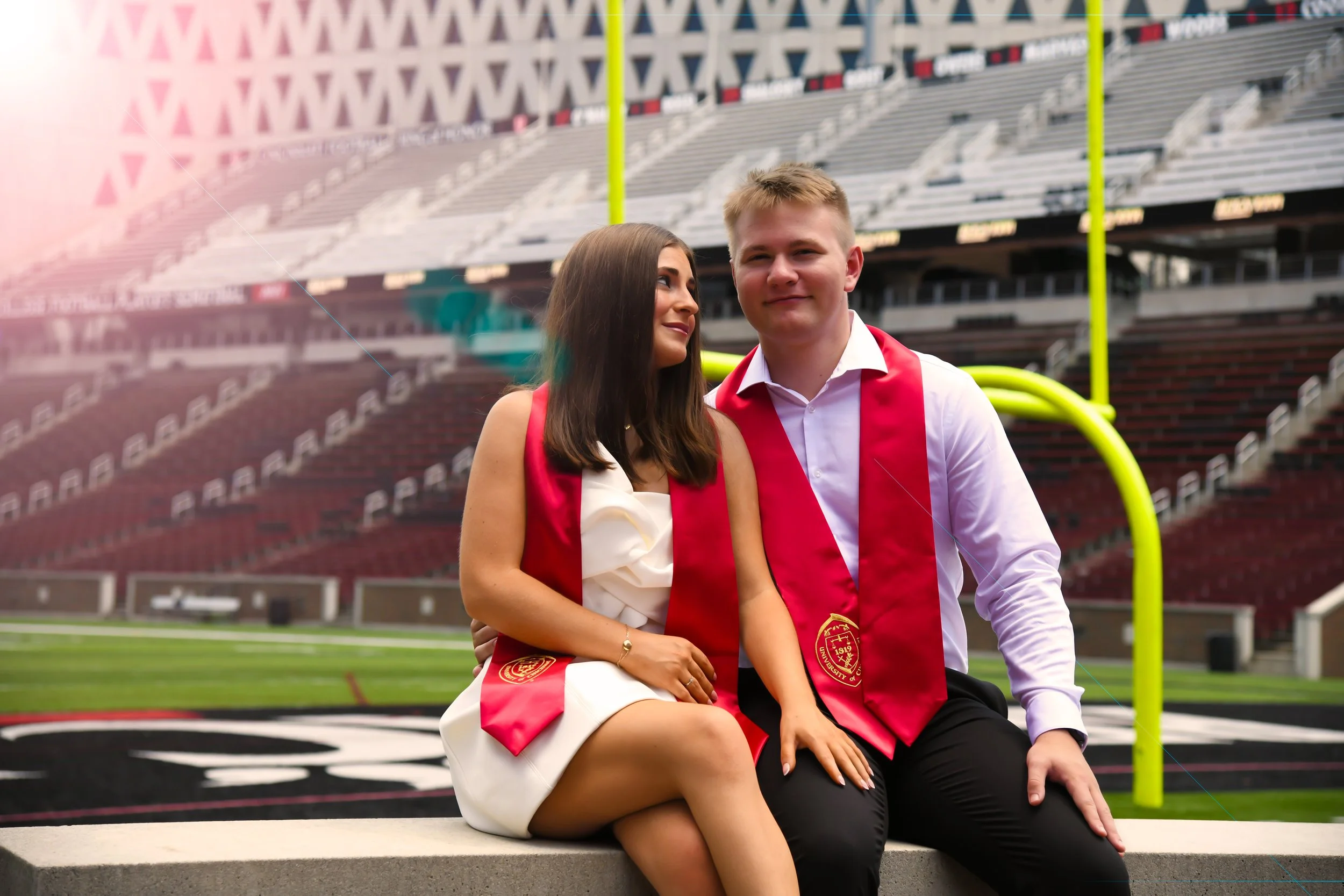 A young man and woman in graduation attire sitting on a bench inside a football stadium, with yellow goalposts and stadium seating in the background.