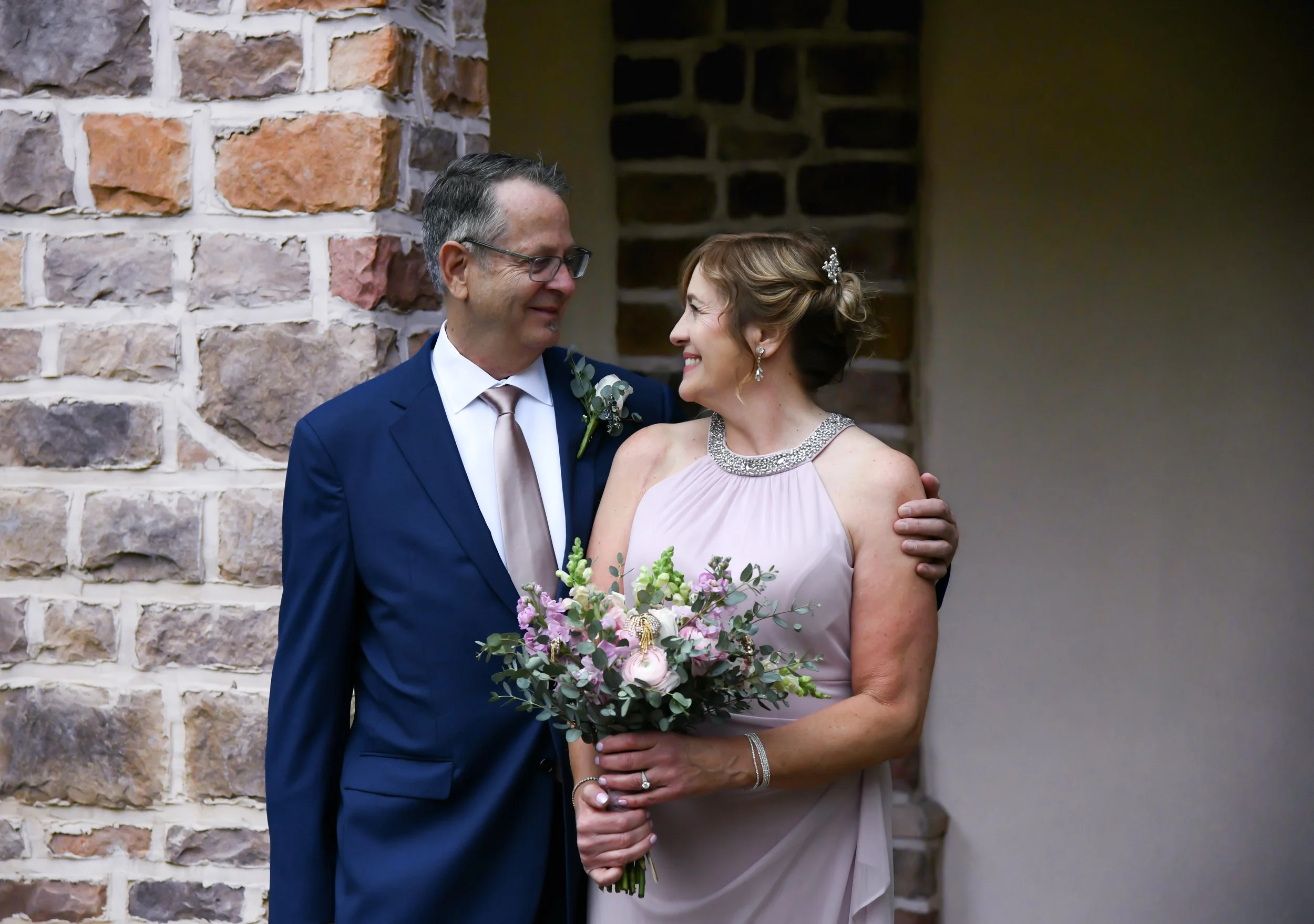 A bride and groom standing close together, smiling at each other during their wedding, with a brick wall in the background.