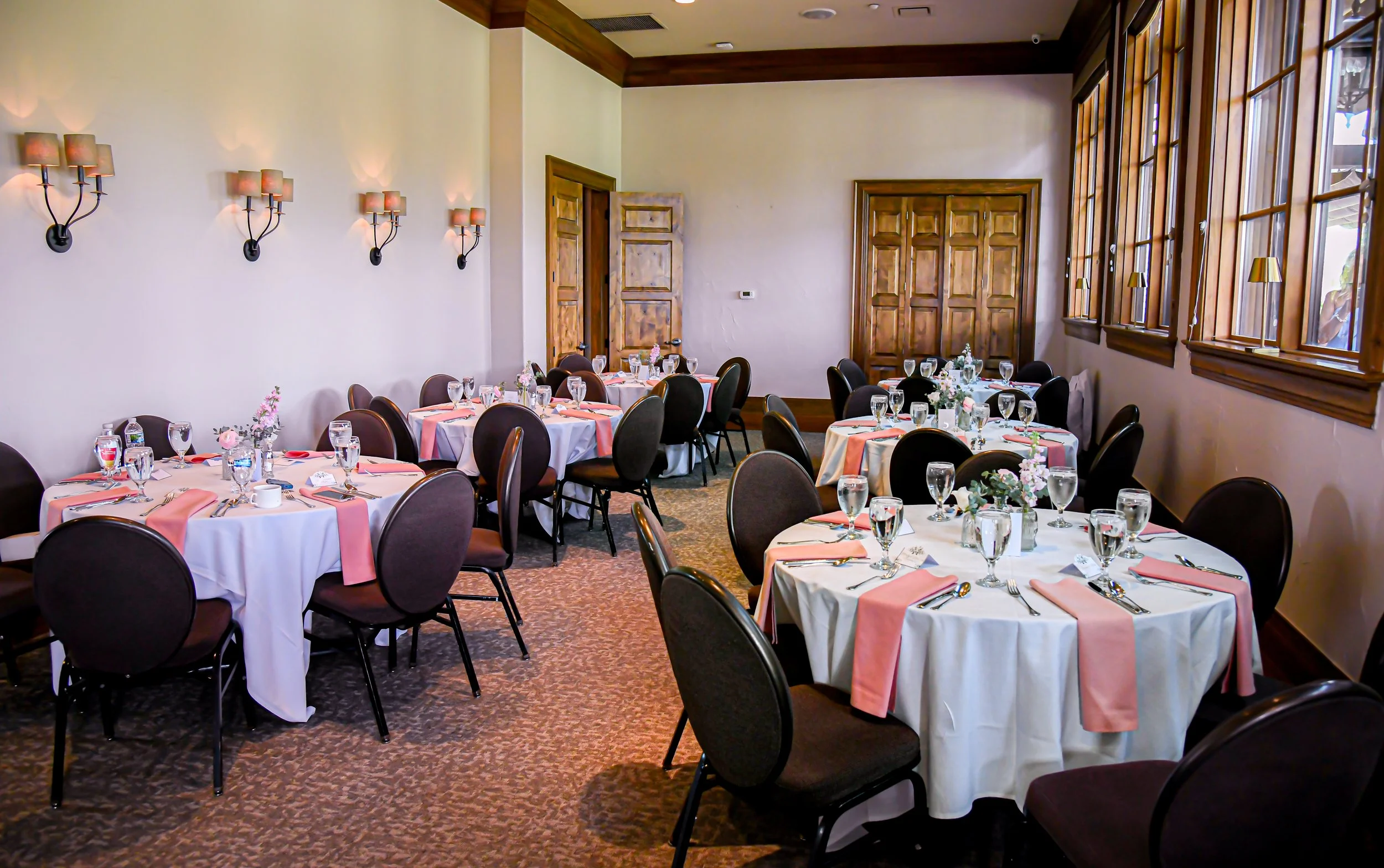 Banquet room set up with round tables covered in white tablecloths, decorated with pink napkins, glassware, silverware, and pink floral centerpieces. The room has cream-colored walls, large wooden windows, and several wall-mounted lamps, with a woode