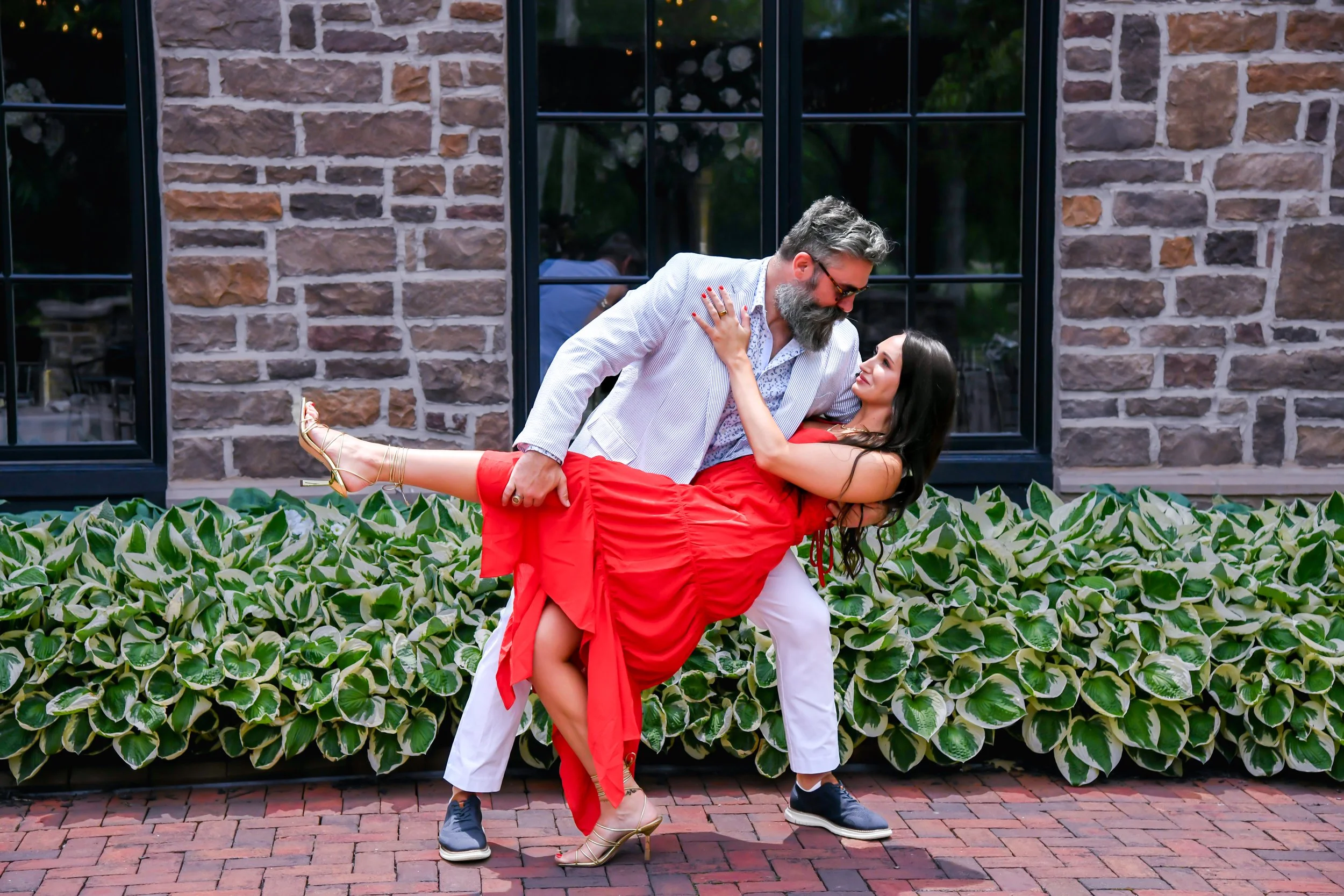 A man with a beard, wearing glasses, a light-colored blazer, and light pants, is holding a woman in a red dress and golden heels in a dance-like pose outside in front of a brick building with large windows and green bushes.