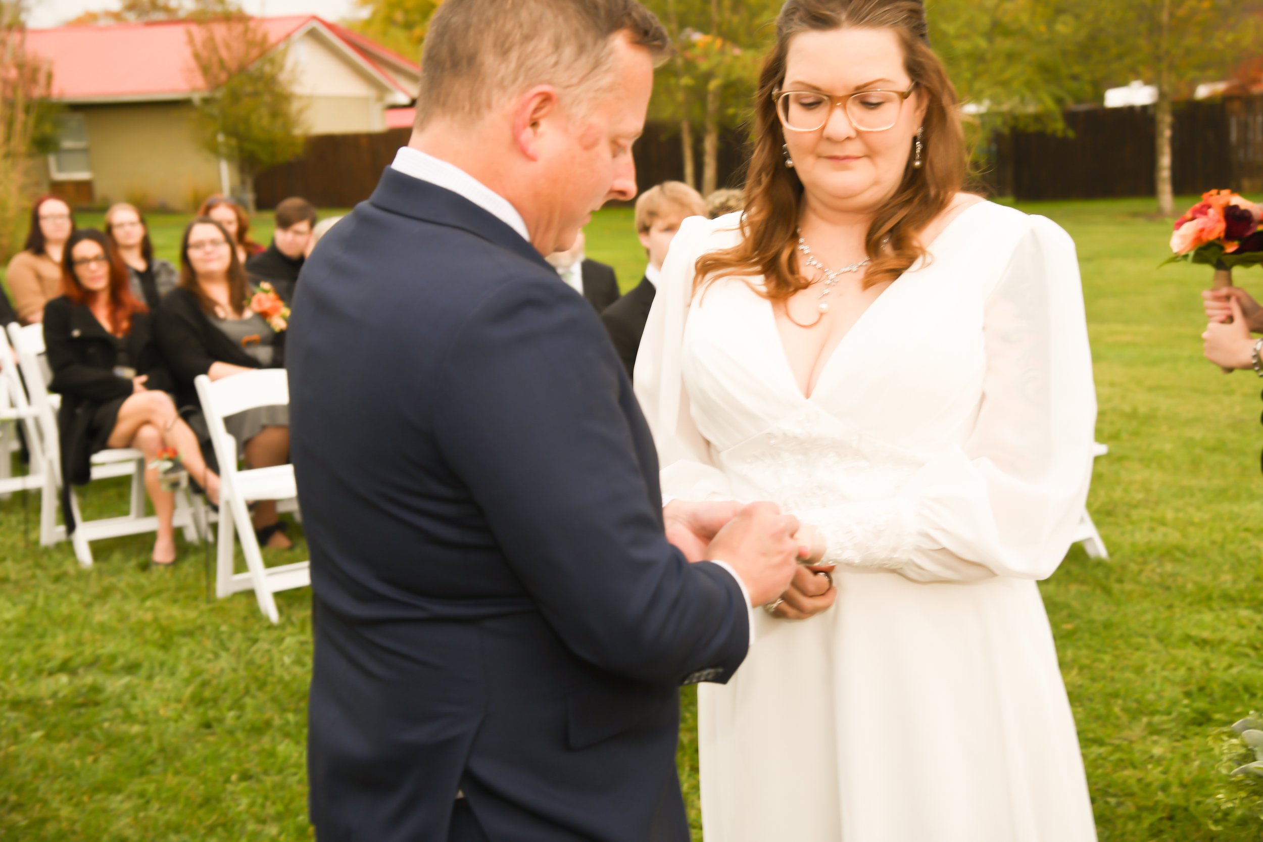 A bride and groom exchange vows during an outdoor wedding ceremony on a grassy lawn. The groom is in a navy suit, and the bride is dressed in a white gown with lace details. Guests are seated on white chairs in the background, watching the ceremony.