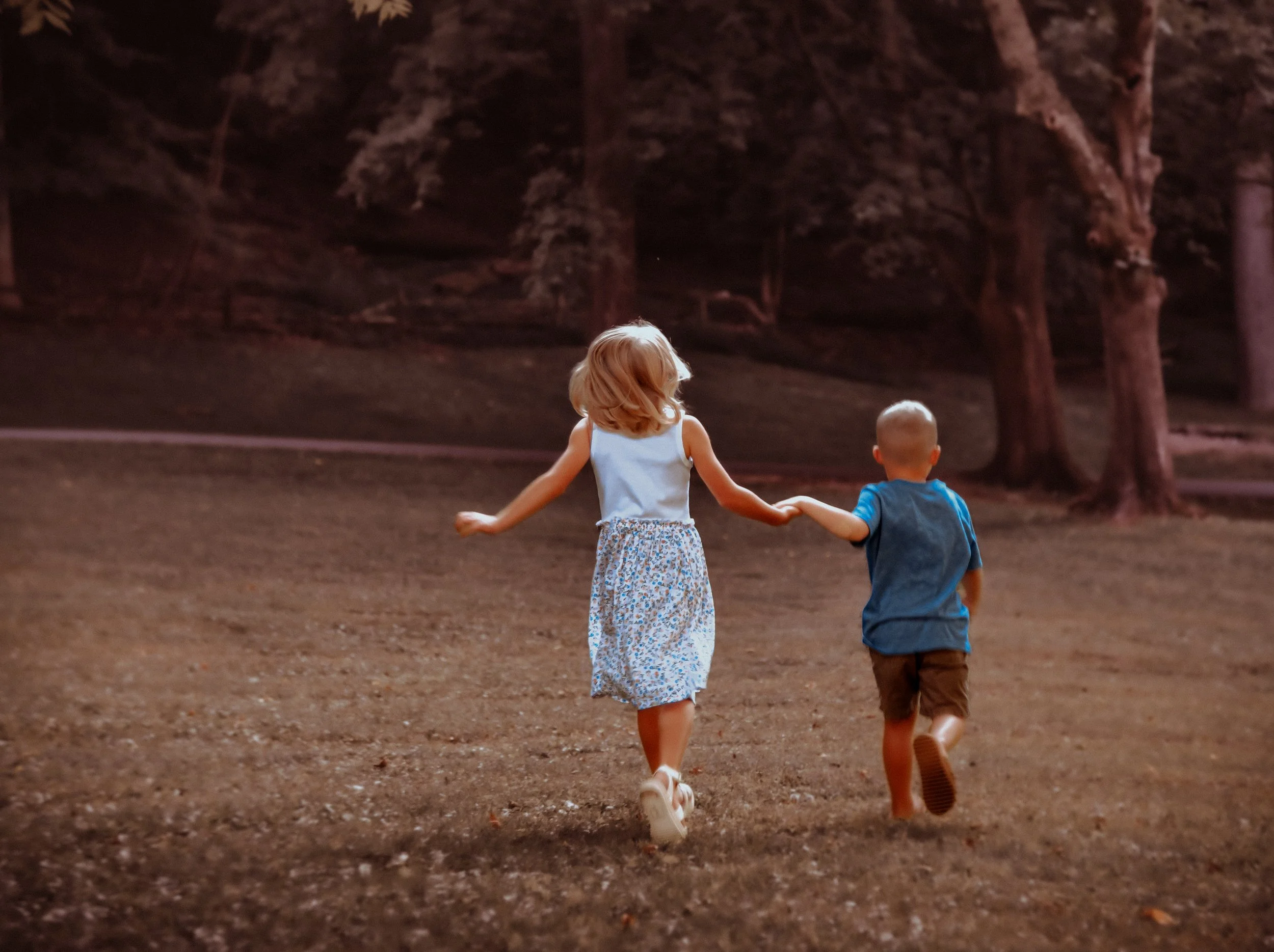 Two children, a girl with light brown hair in a white top and floral skirt and a boy with a shaved head in a blue shirt and brown shorts, holding hands and running on a dirt path through a wooded area.
