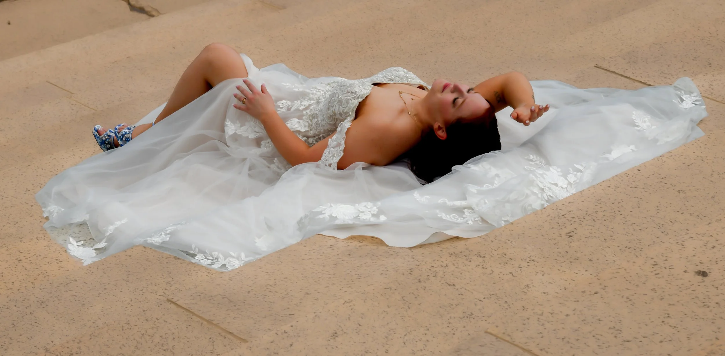 A woman in a white wedding dress lying on her back on a beige tiled floor, eyes closed, one hand resting on her stomach, the other arm bent with her hand near her face.