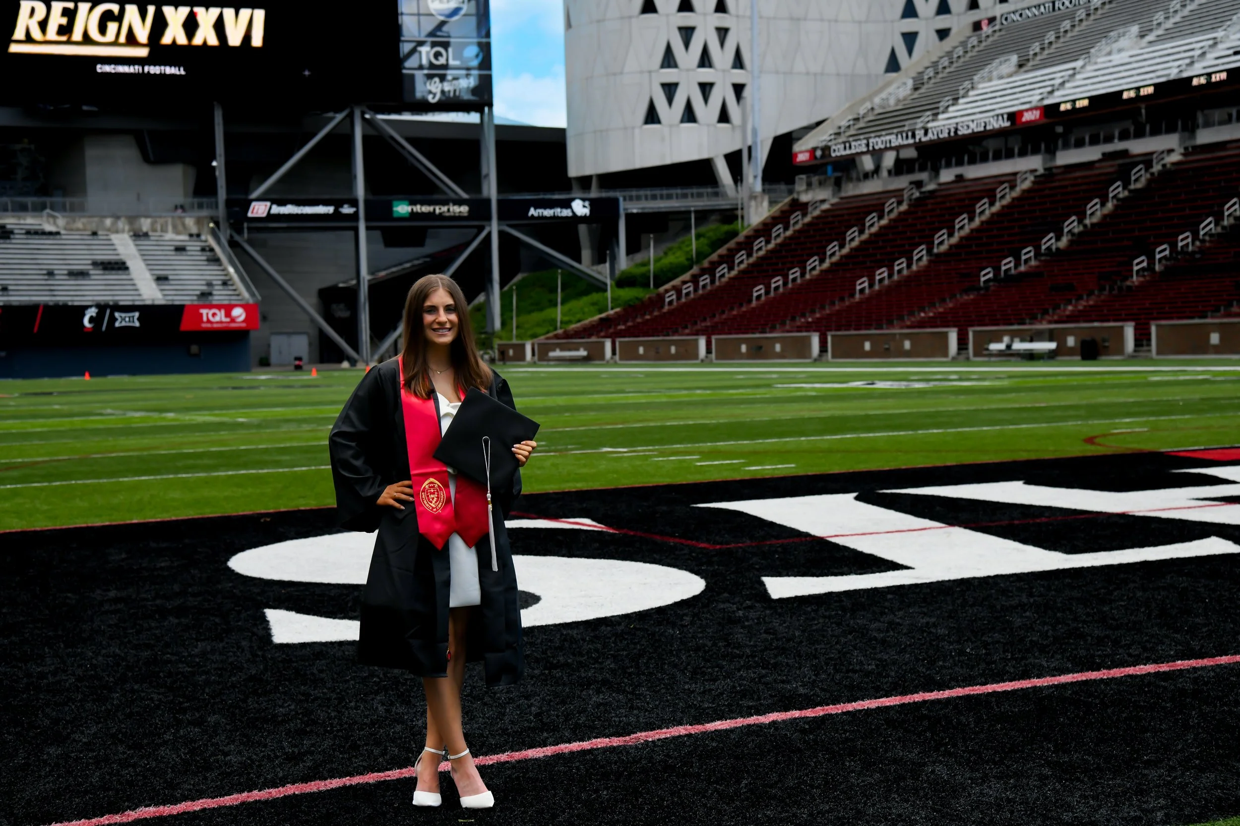 A young woman in a graduation gown standing on a football field at a stadium with empty seats, holding a diploma and smiling.