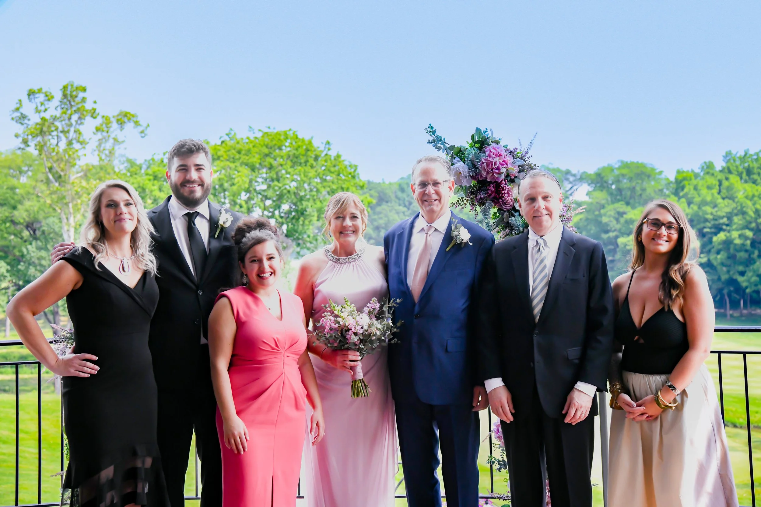 Group of eight people standing outdoors, dressed in formal attire, with a background of trees and a body of water, at a wedding or celebration event.
