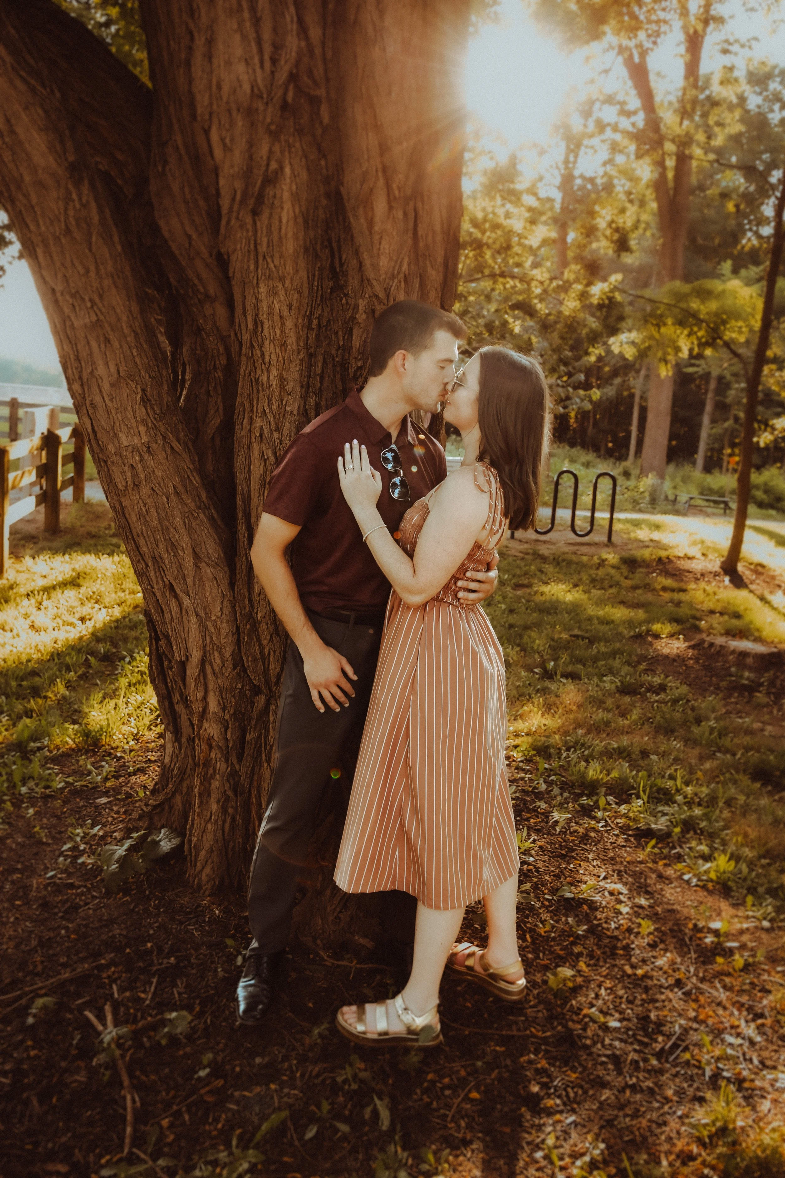 A young couple shares a kiss while standing against a large tree in a park during sunset.