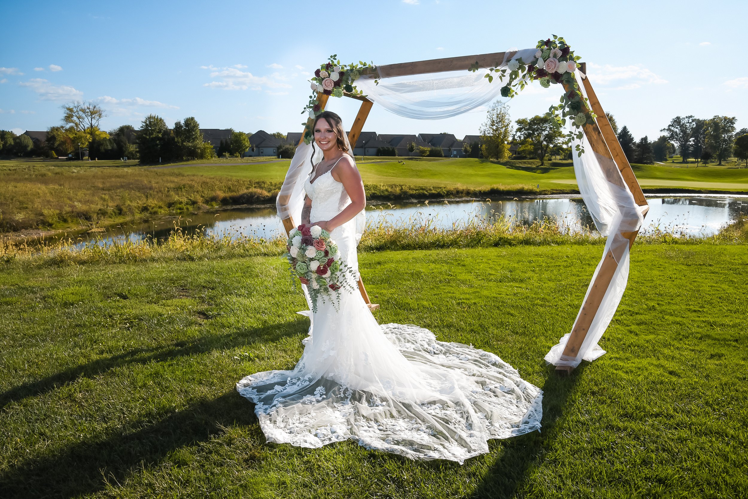 A bride in a white wedding dress holding a bouquet of flowers, standing outdoors under a decorated wooden arch near a pond, with green grass, trees, and houses in the background on a sunny day.