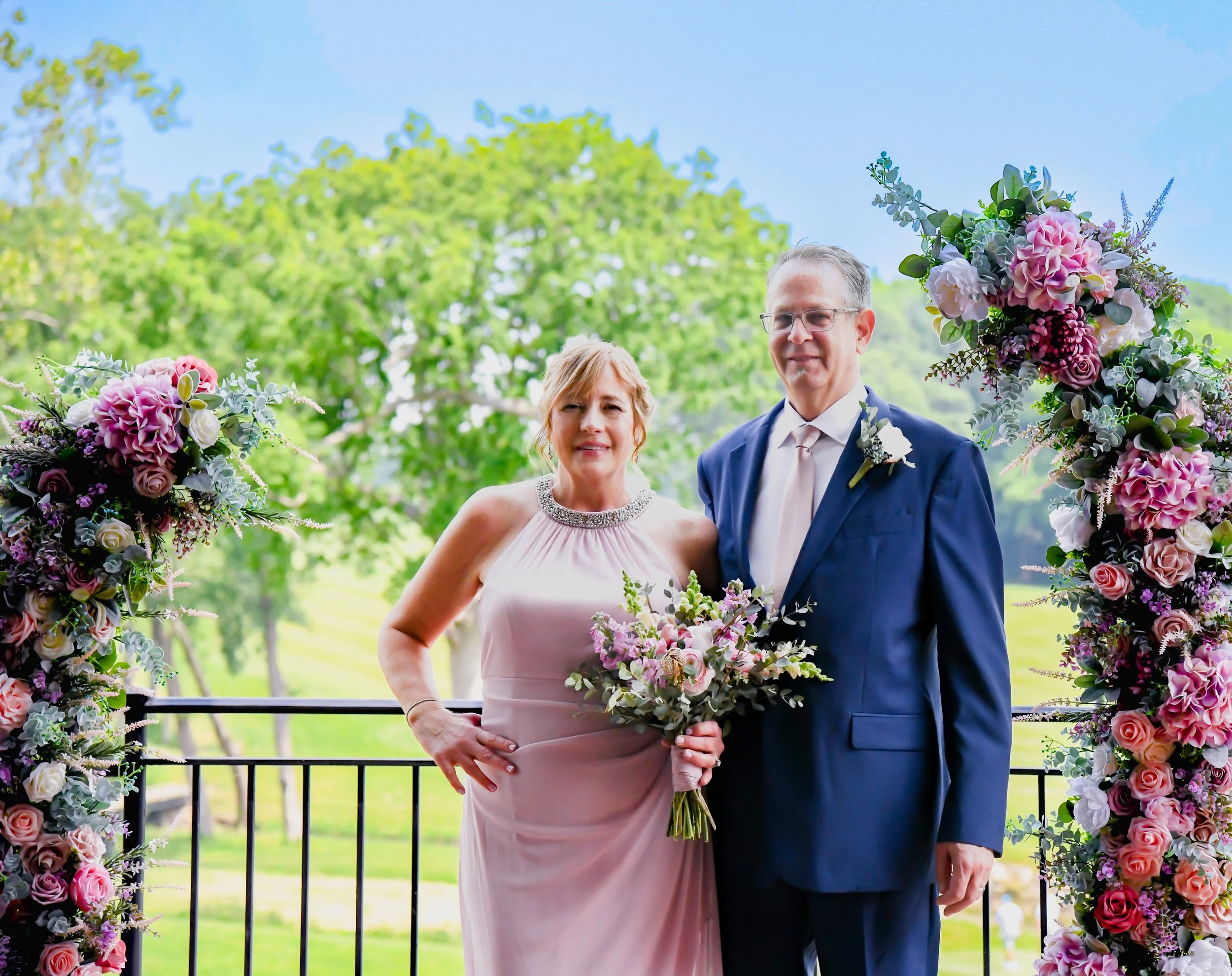 A woman in a pink dress and a man in a blue suit with a white shirt standing outside in front of a floral arch with pink and white flowers, holding a bouquet and smiling, with green trees and a blue sky in the background.