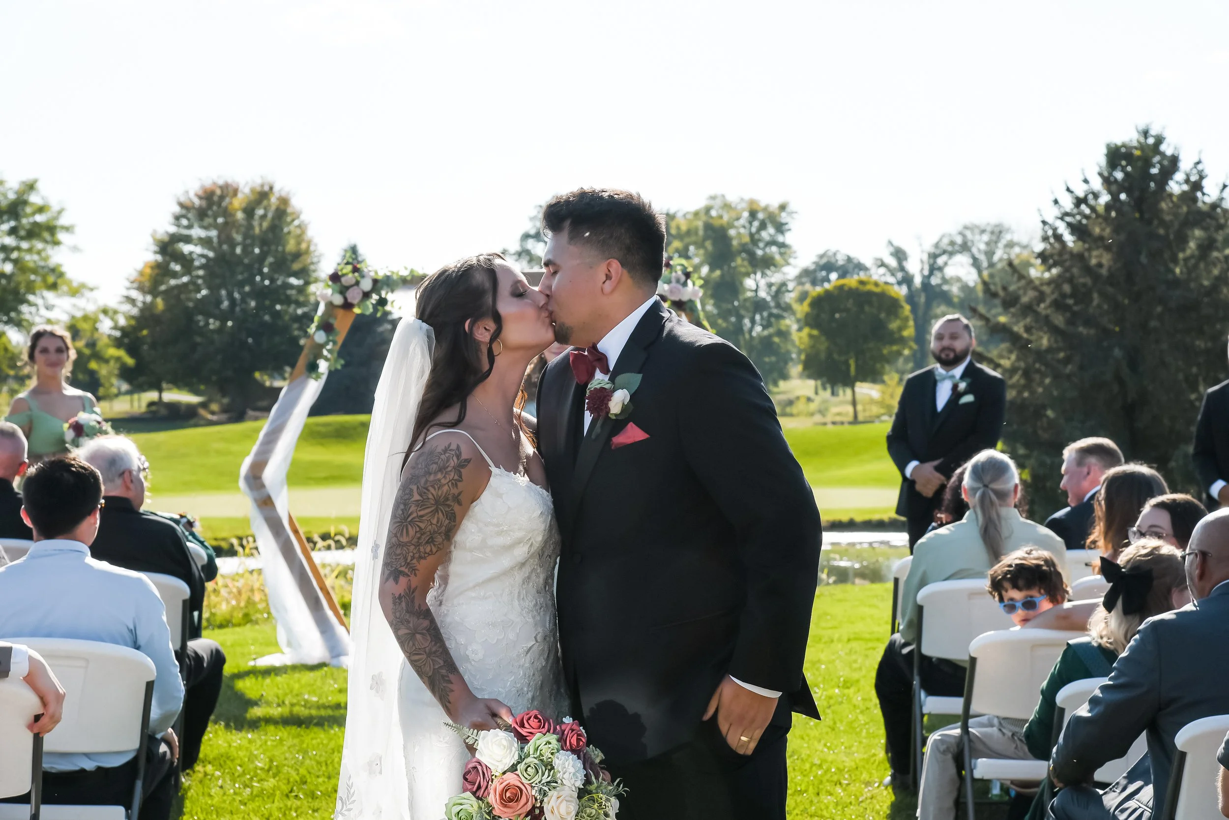A wedding ceremony outdoors with a bride and groom kissing in front of friends and family on a sunny day.