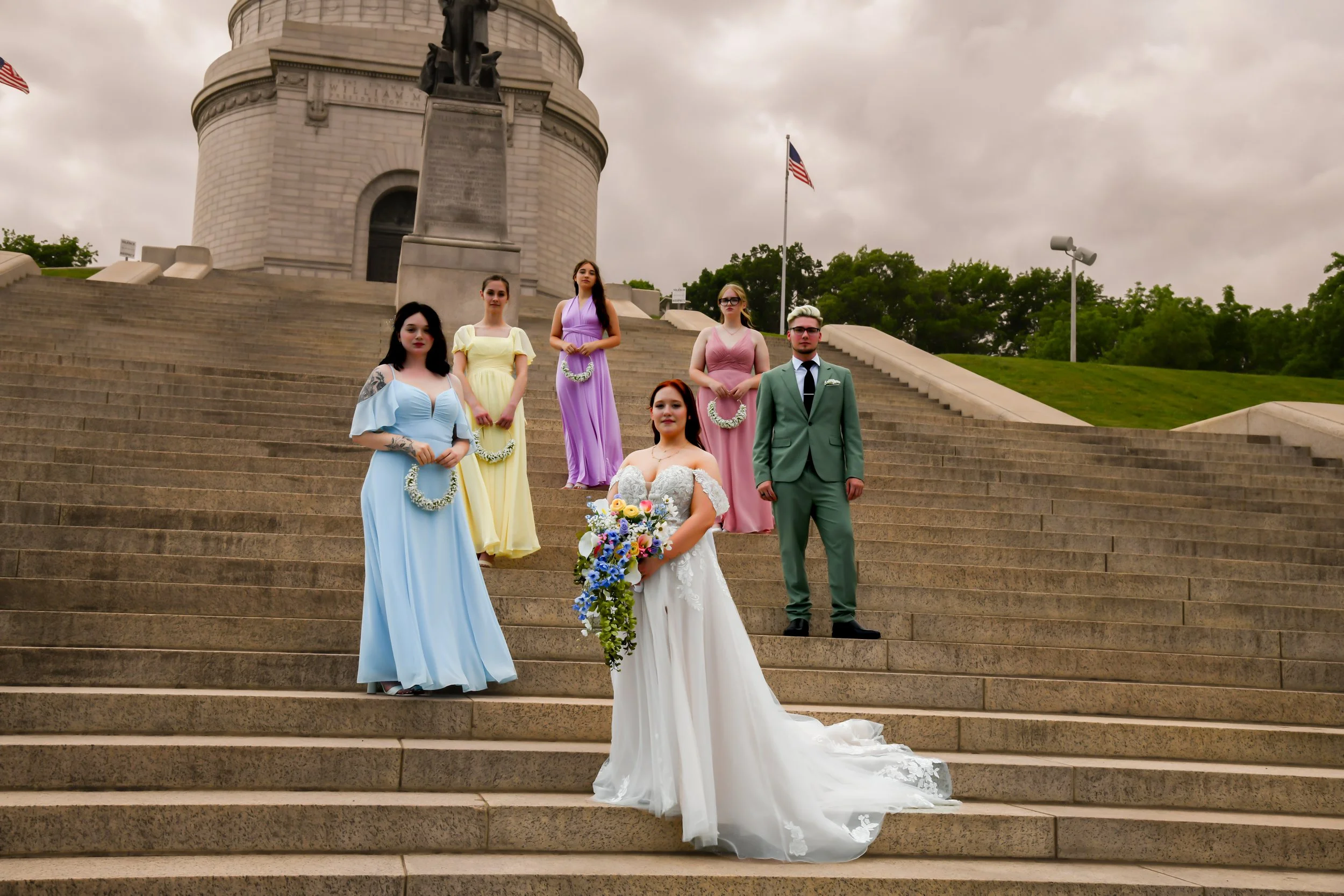 A bride in a white wedding dress holding a bouquet of colorful flowers stands on stairs in front of five bridesmaids and one groomsman in pastel-colored dresses and a suit at a monument with stairs, a flag, and trees surrounding.