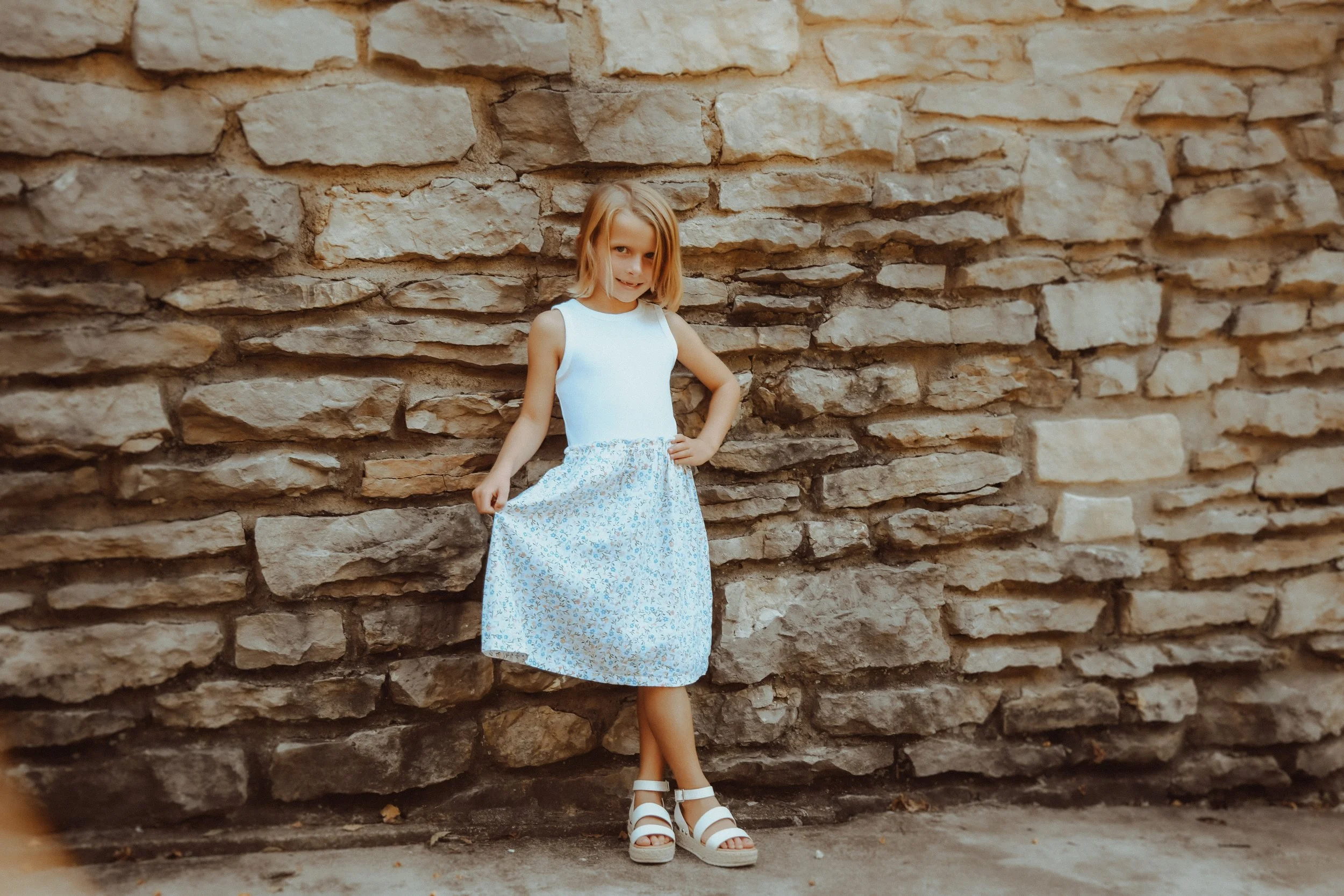 A young girl with blonde hair wearing a white sleeveless top, a blue floral skirt, and white sandals standing against a textured stone wall, posing with one hand on her hip.