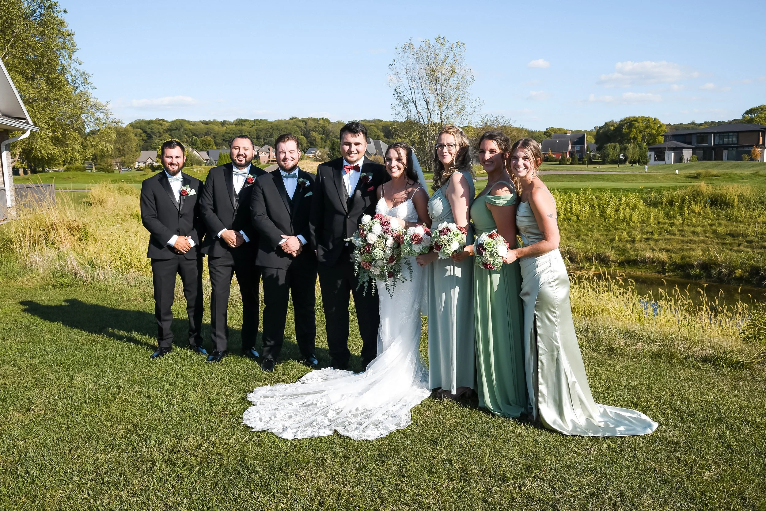 A wedding party of eight people standing outdoors on a sunny day near a pond, dressed in formal attire with the bride and groom in the center, surrounded by bridesmaids in green dresses and groomsmen in black tuxedos.