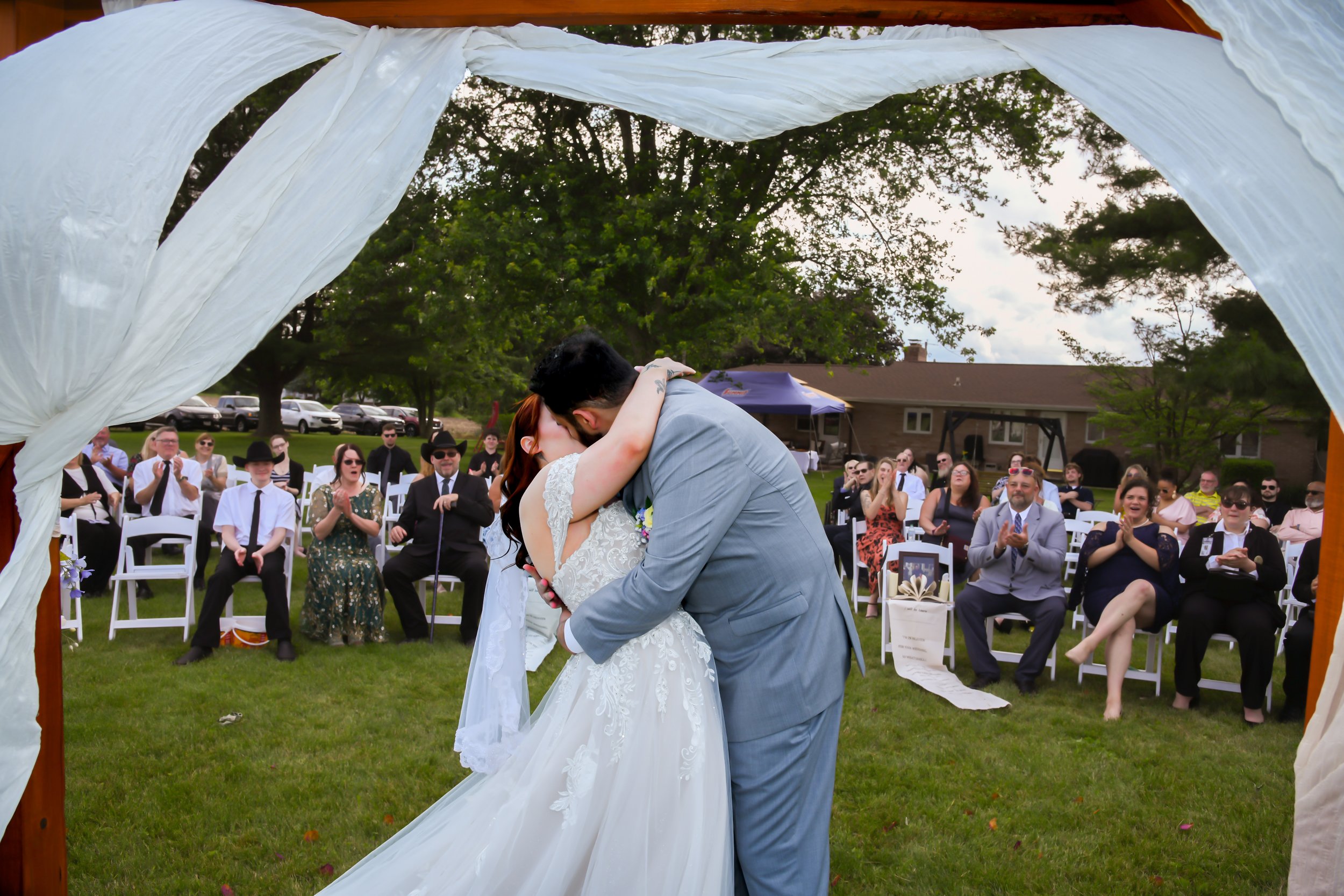 A bride and groom share a kiss during their outdoor wedding ceremony, surrounded by seated guests under a wooden arch with white fabric decorations, with trees and a house in the background.