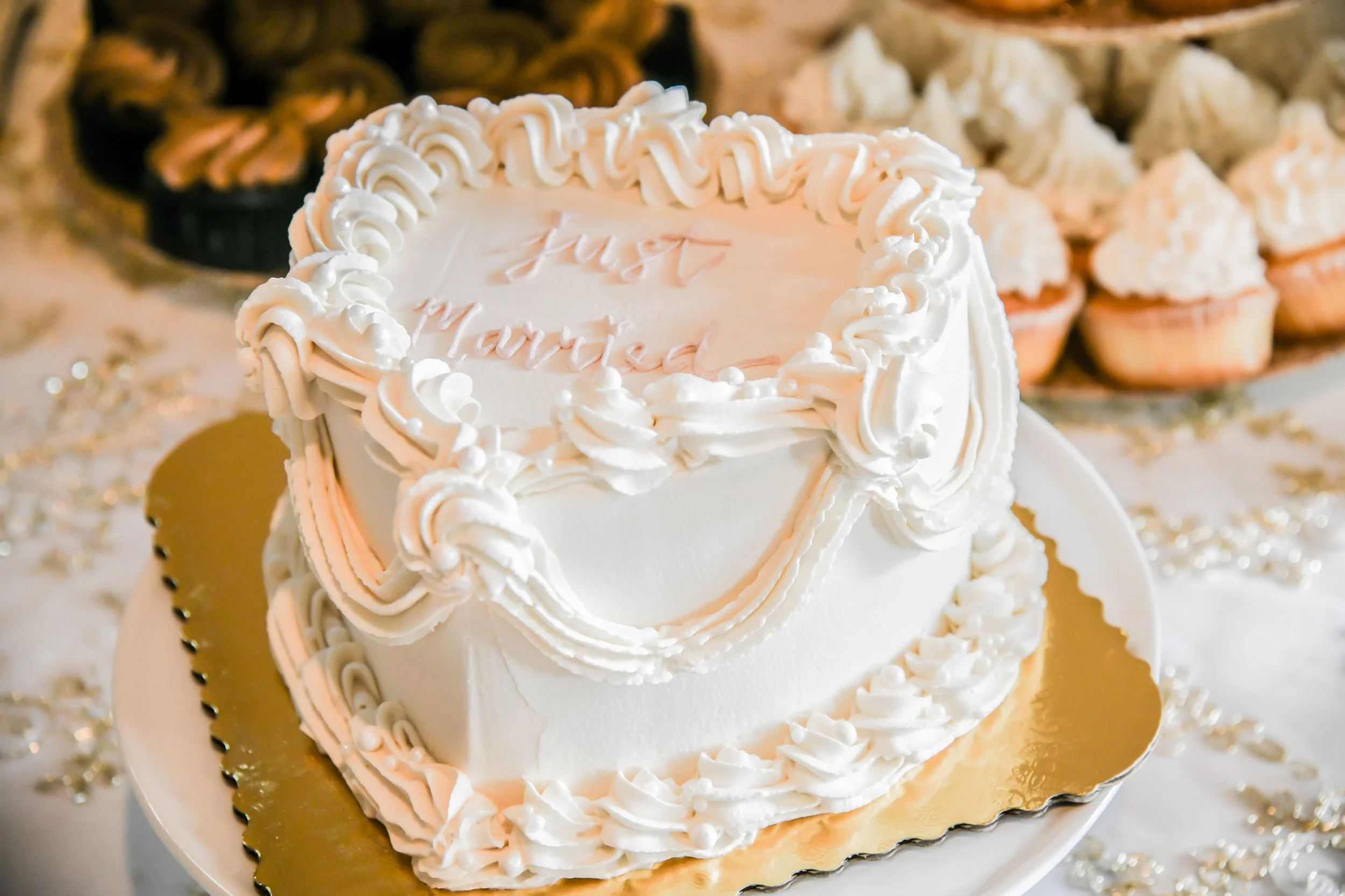 A white heart-shaped birthday cake with pink and white frosting decorations and a message that says "Just Married" written in pink on top, surrounded by cupcakes and decorative items.