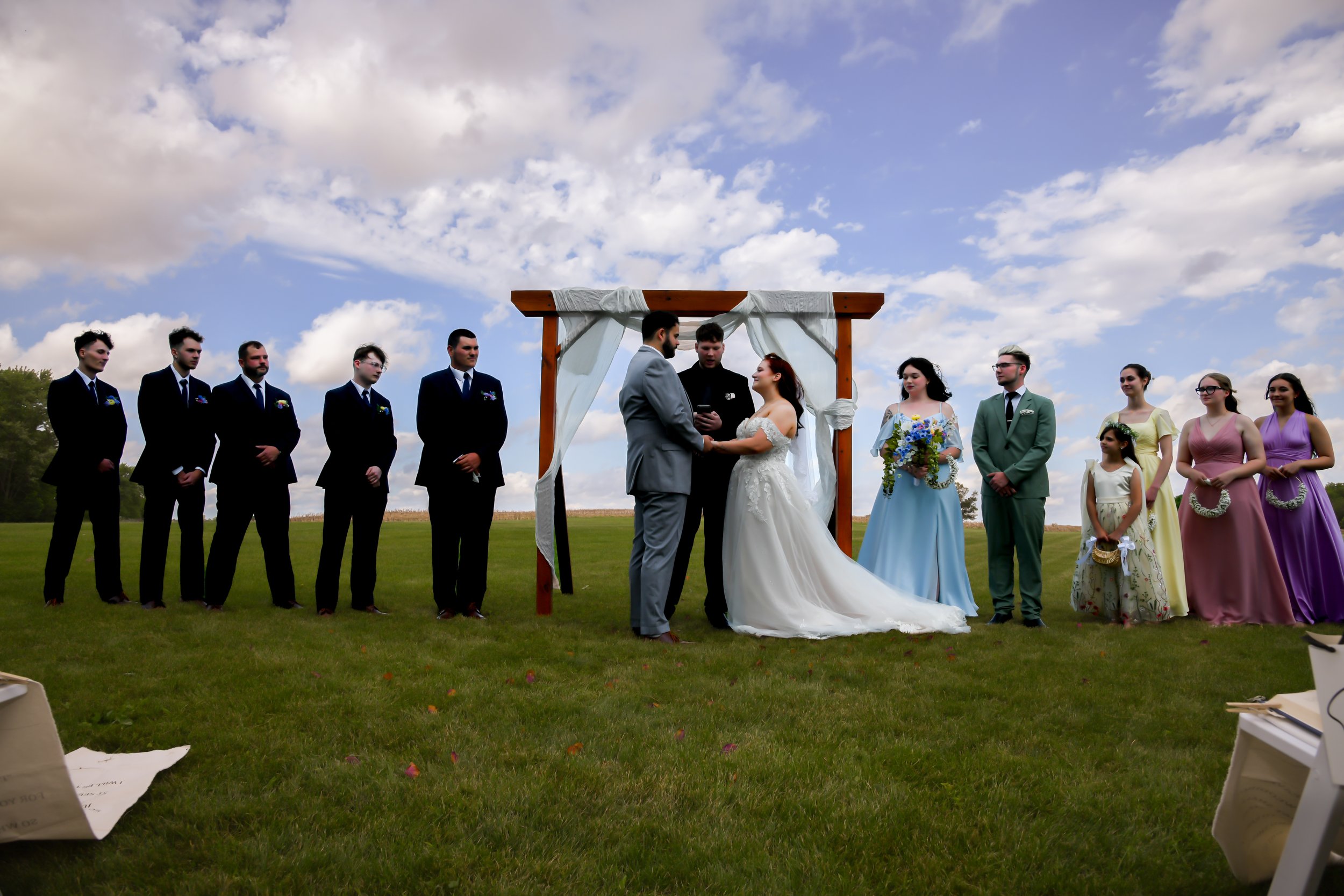 A group of people participating in a wedding ceremony outdoors under a wooden arch with white fabric, with a bride and groom exchanging vows, surrounded by bridesmaids and groomsmen, on a grassy field under a partly cloudy sky.