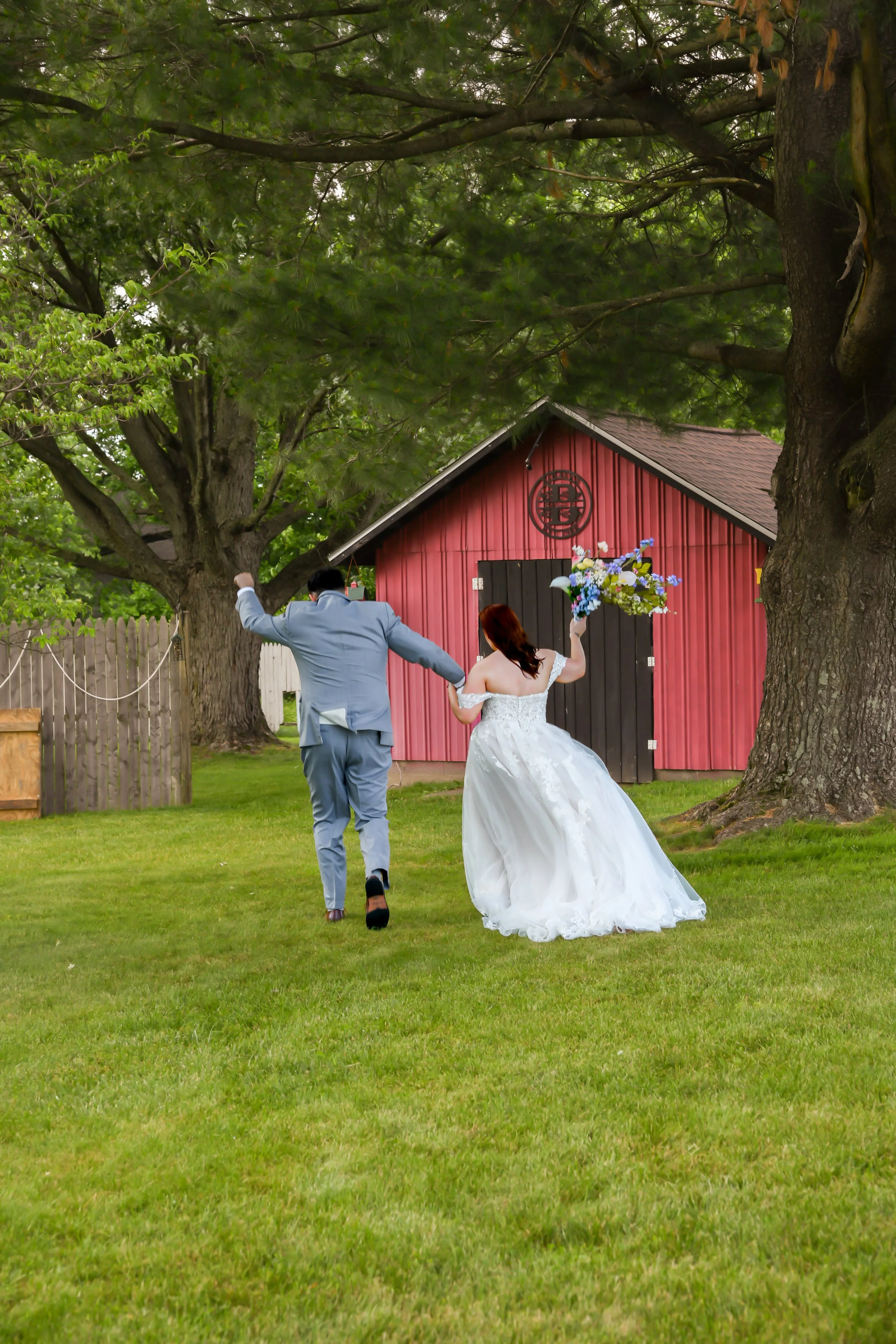 A bride in a white wedding dress holding a bouquet and a groom in a blue suit running towards a red barn with a peace symbol and large trees in the background.