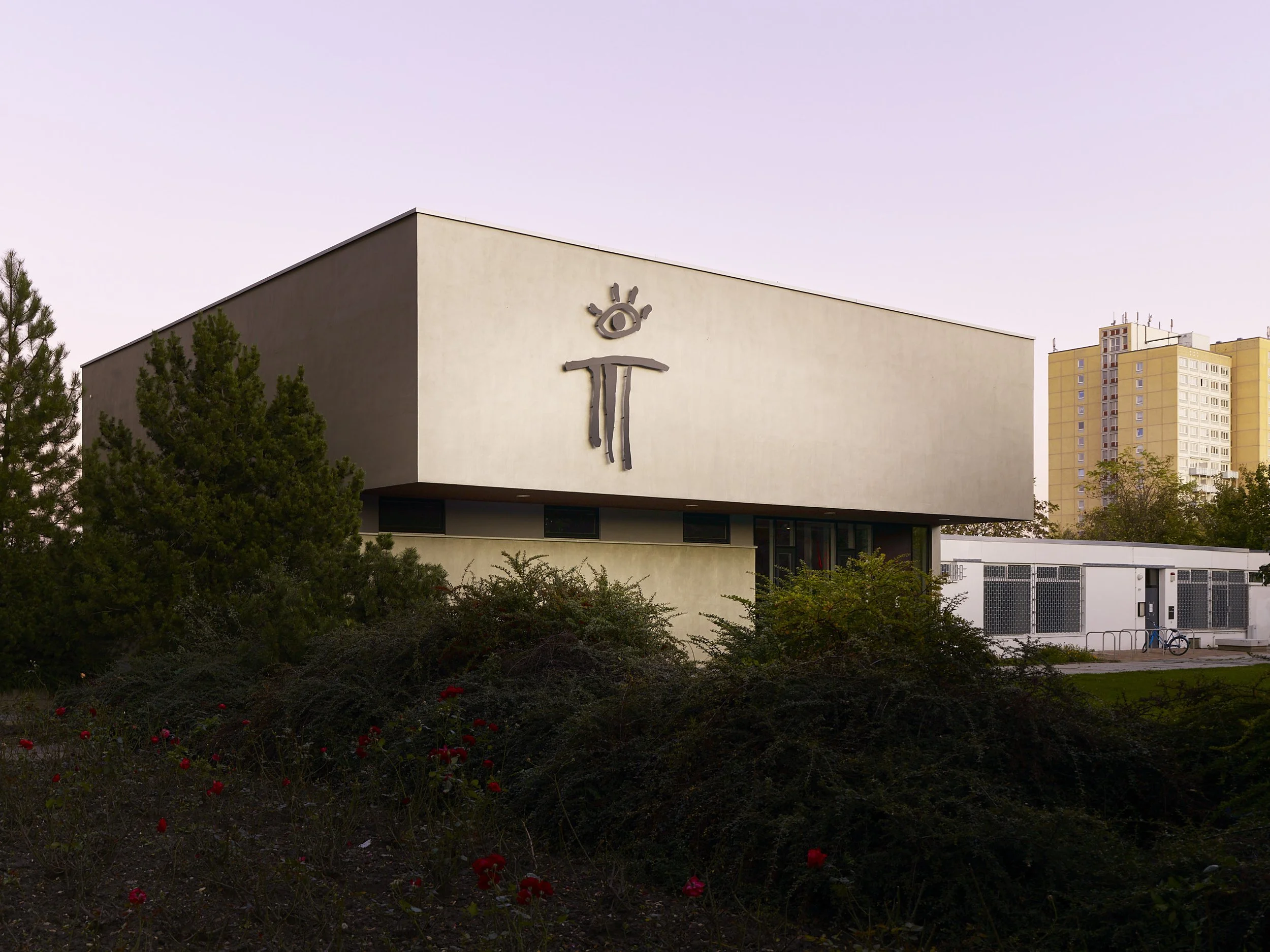 Modernes Gebäude mit einer großen Skulptur an der Wand, umgeben von Büschen und Bäumen, mit hohen Wohngebäuden im Hintergrund.