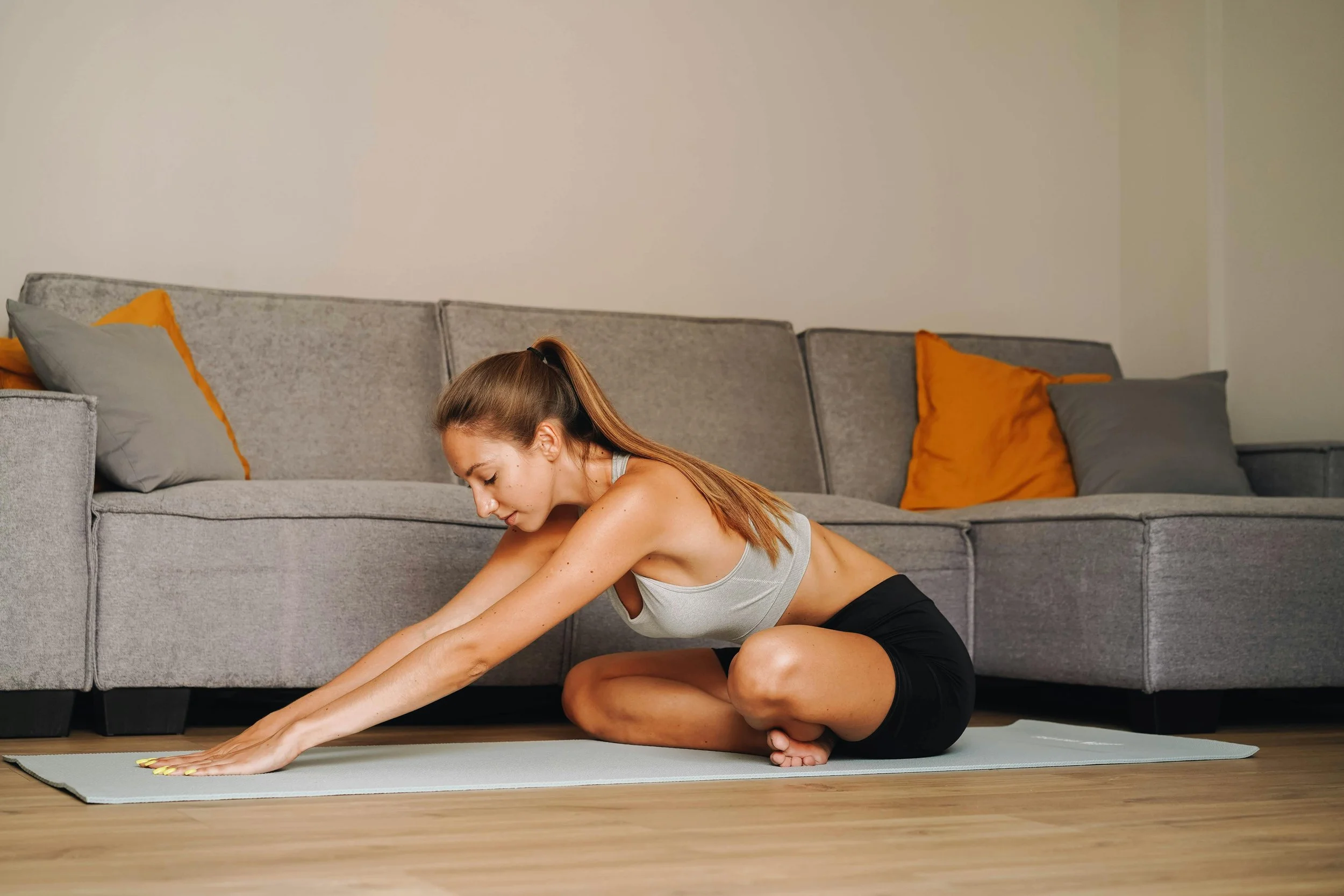 The image shows a person performing a stretching exercise on a light coloured Pilates mat placed on a wooden floor. The person is sitting with one leg folded under their body and the other extended forward reaching out with both arms.