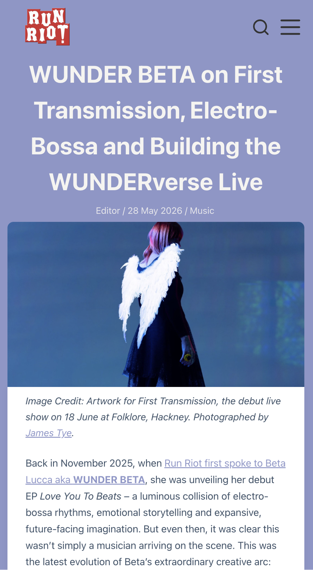 WUNDER BETA (Roberta Lucca) dressed in a black outfit with white angel wings, standing against a blue background.