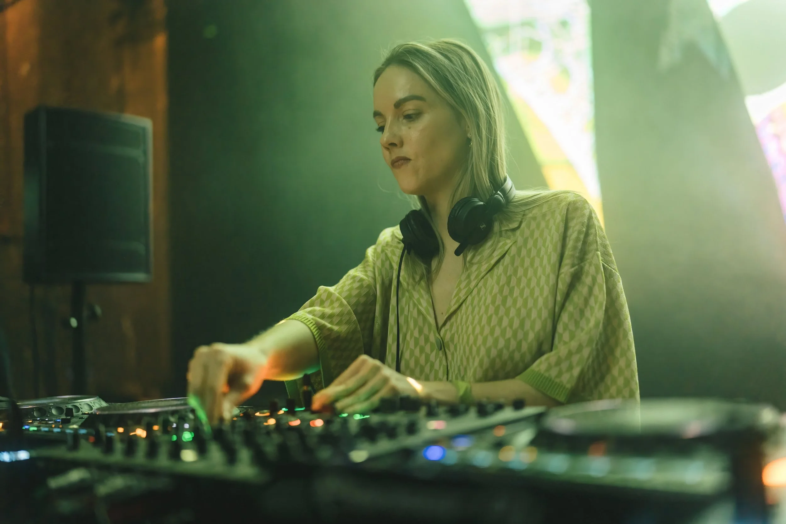 A female DJ wearing headphones adjusts controls on a DJ mixer in a dimly lit venue with green lighting.