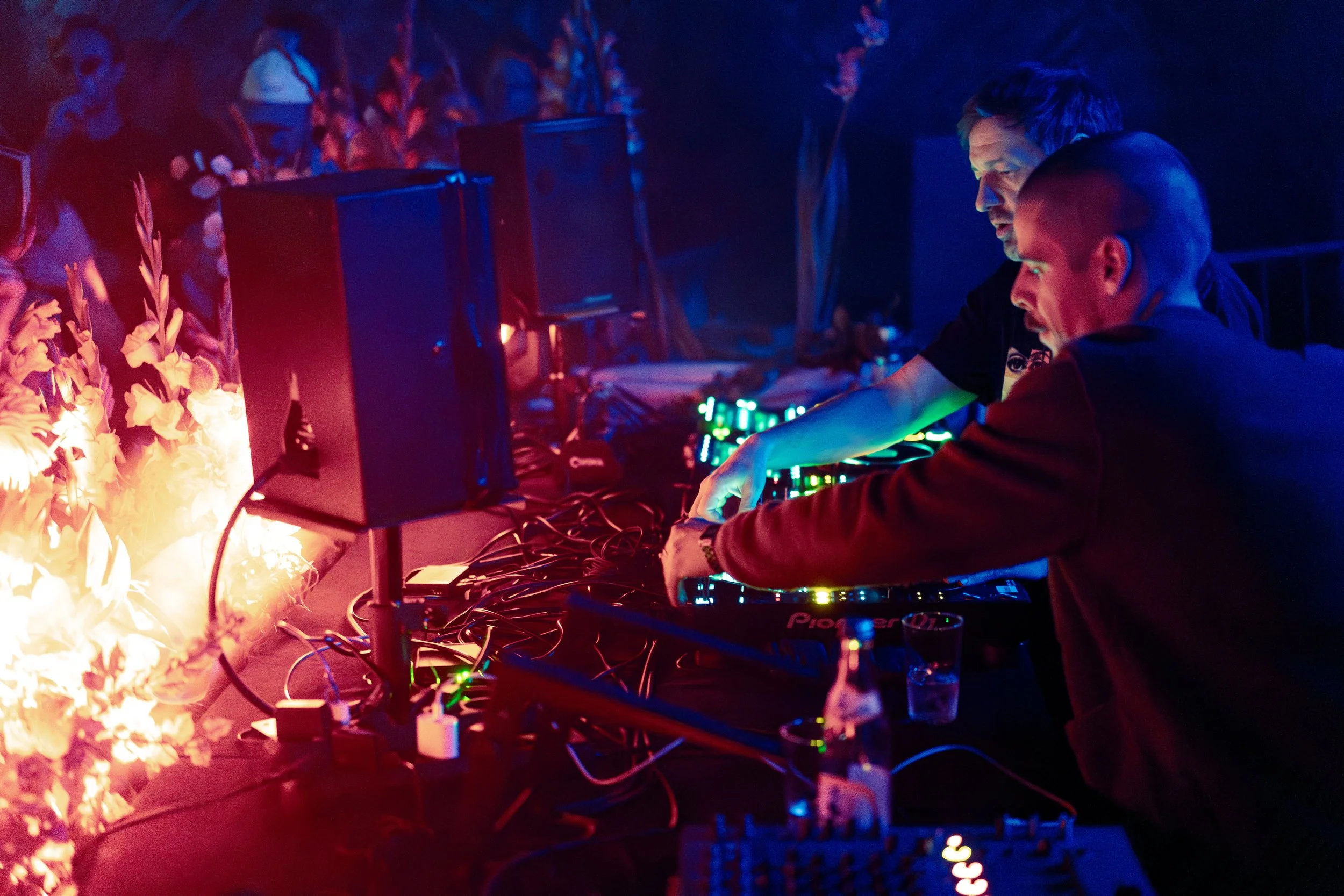 Two men DJing at a club with colorful lights and flames nearby.