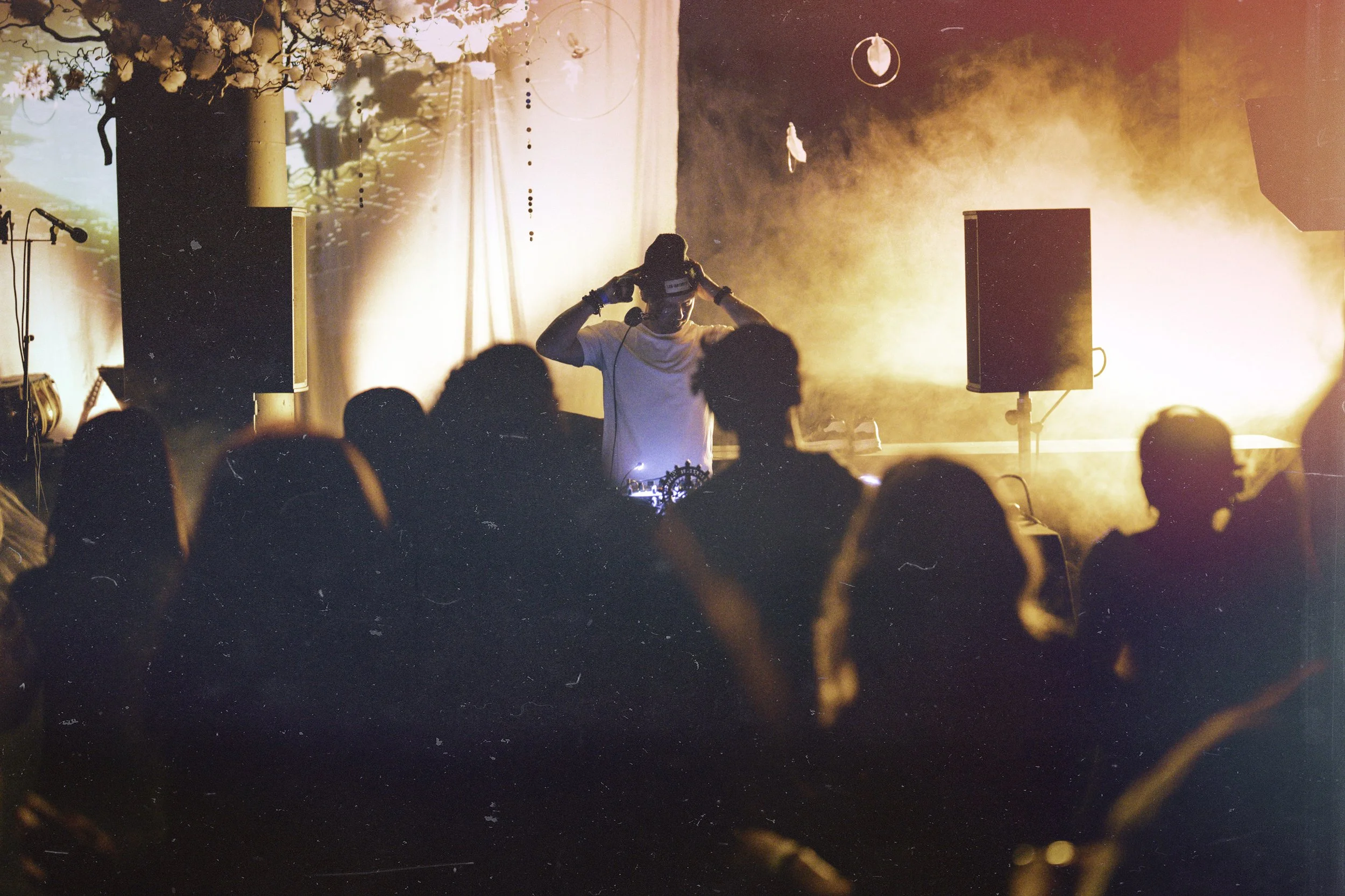A DJ performing on stage with bright orange lighting, fog, and speakers, while a crowd of people watches at an indoor music event.