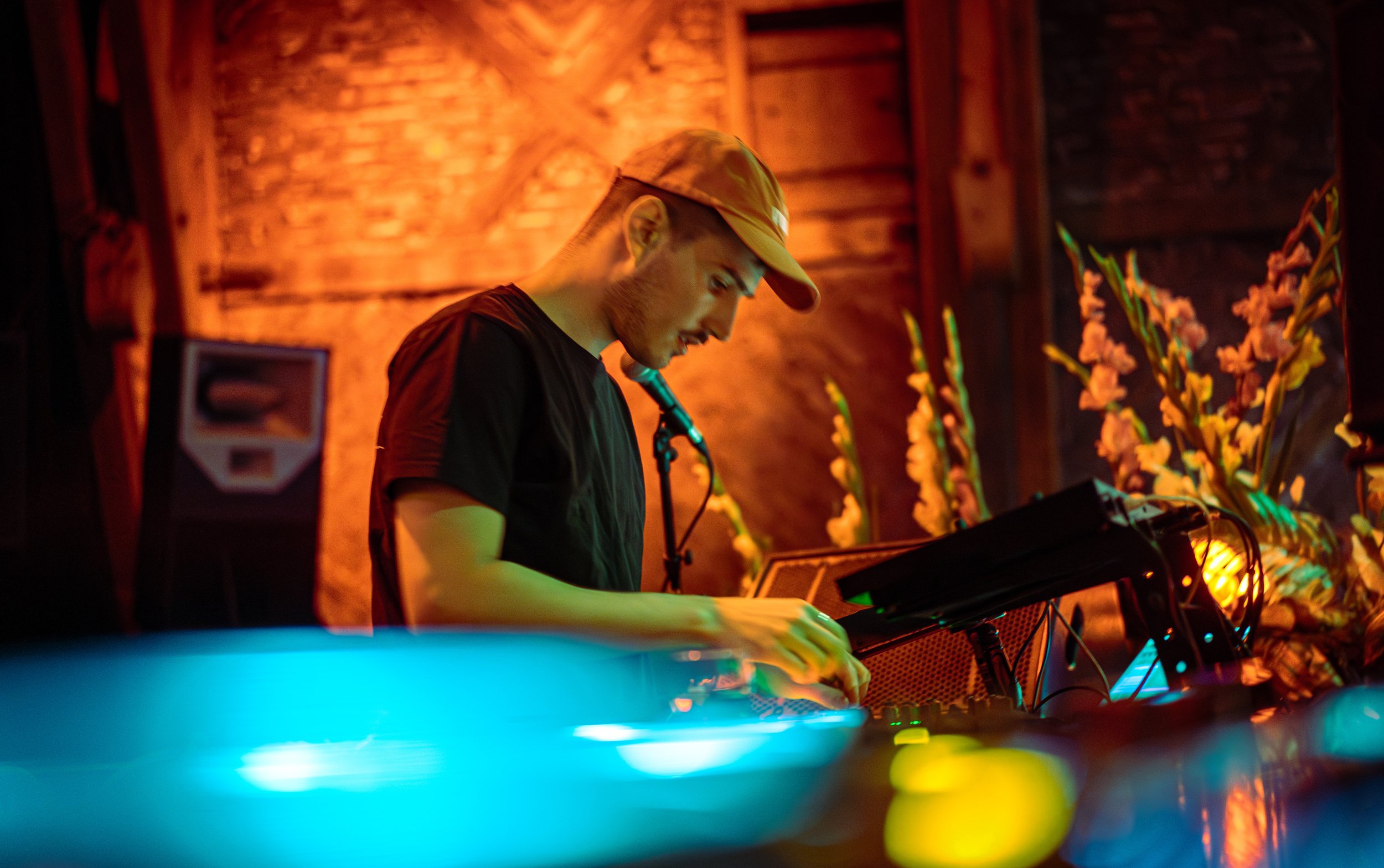 A DJ in a black shirt and beige cap operating sound equipment with colorful lighting and flowers in the background.