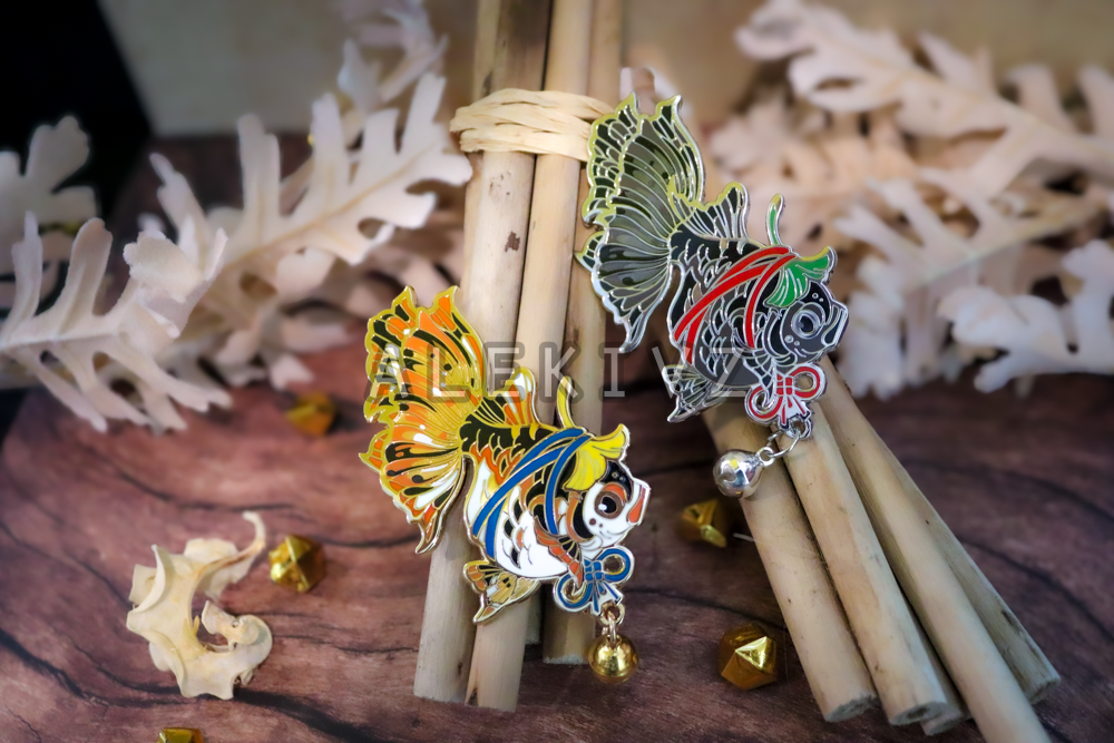 Example of enamel pins for sale, featuring a goldfish with a ginkgo leaf tied to its head like an adornment. Features multiple layers of enamel and screenprinting as well as an attached bell.