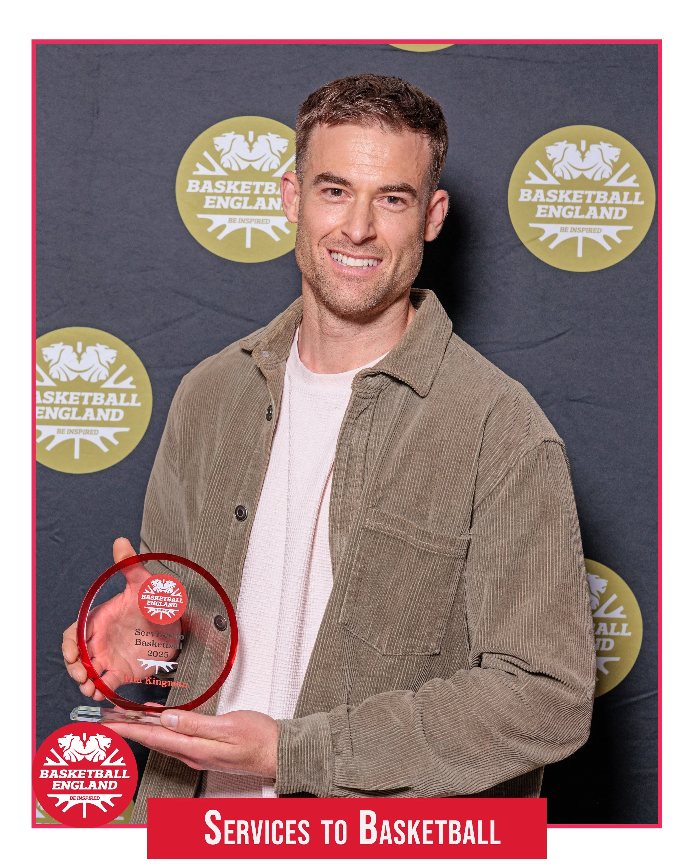 A smiling man holding a circular red glass award in front of a dark backdrop patterned with gold Basketball England logos. He is wearing a white shirt and a green corduroy jacket, and a red banner at the bottom reads "Services to Basketball"