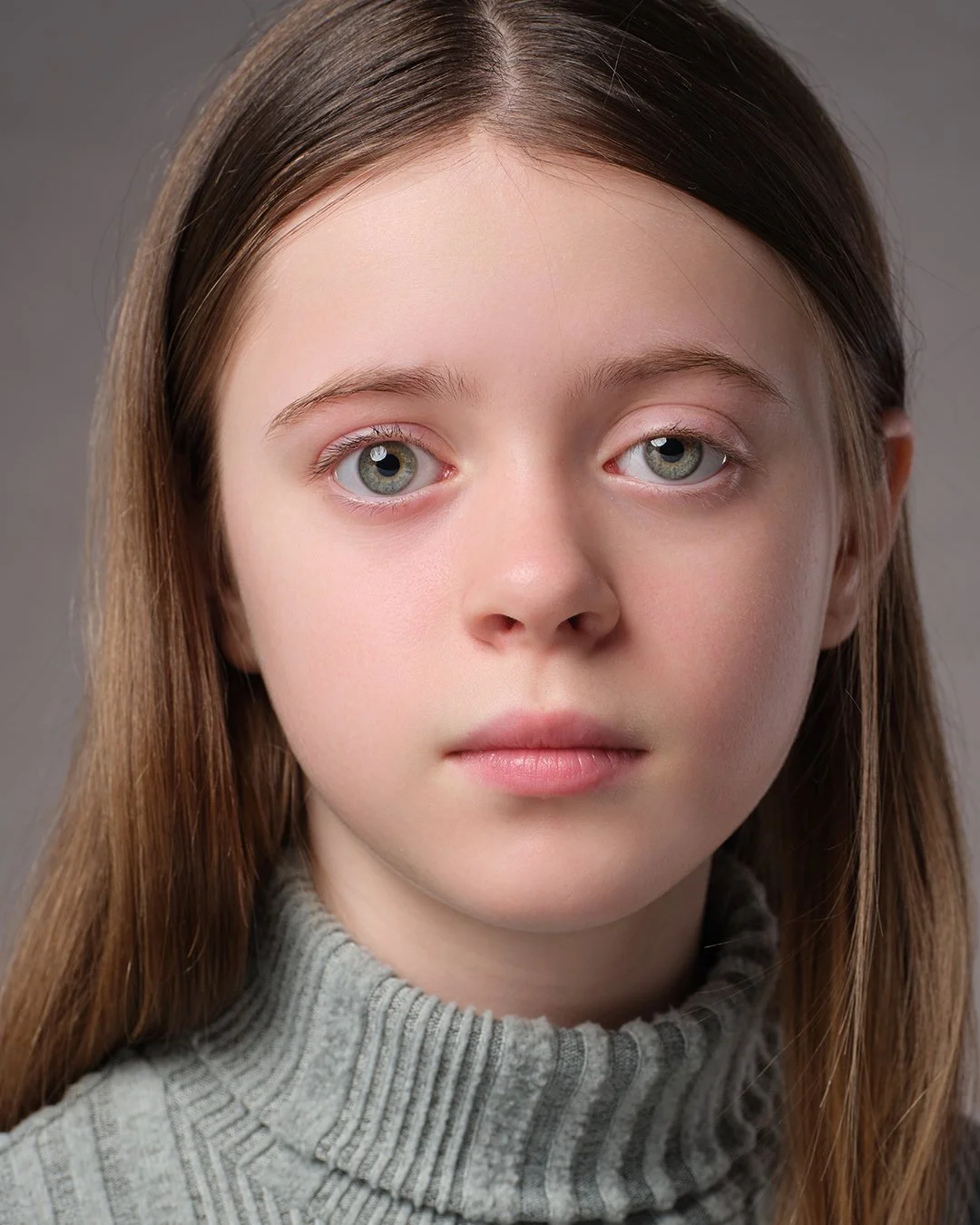 
A close-up portrait of a young person with long, straight light brown hair parted down the middle. They have light-colored eyes and a neutral expression, looking directly at the camera. They are wearing a grey ribbed turtleneck sweater against a sol
