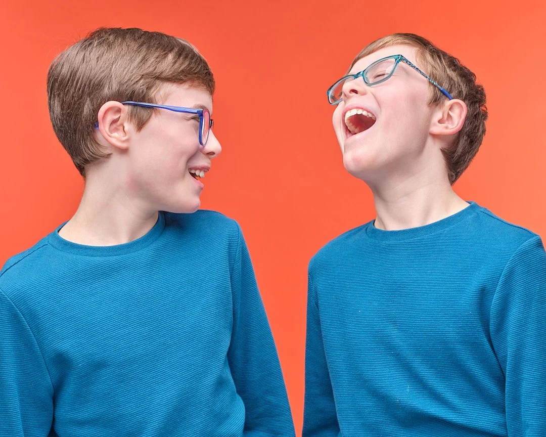 Professional studio headshot of a young boy with glasses laughing, captured against a vibrant orange background. This high-energy child actor headshot demonstrates natural expression and personality for talent agency submissions.