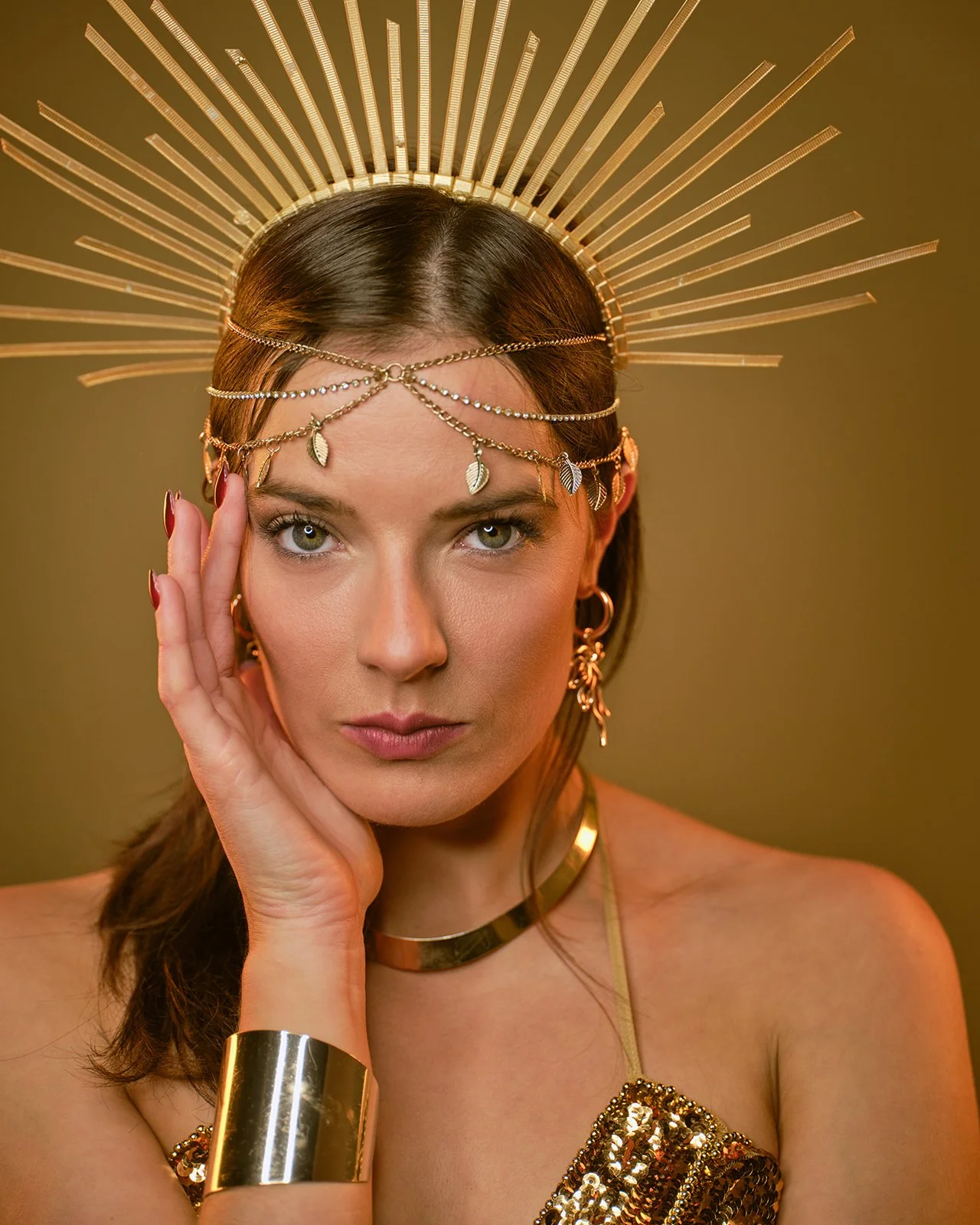 Close up studio portrait of a woman wearing a sunburst gold crown, delicate forehead chain with leaf charms, thick gold cuff bracelet, and a sequined top against a warm gold studio background.