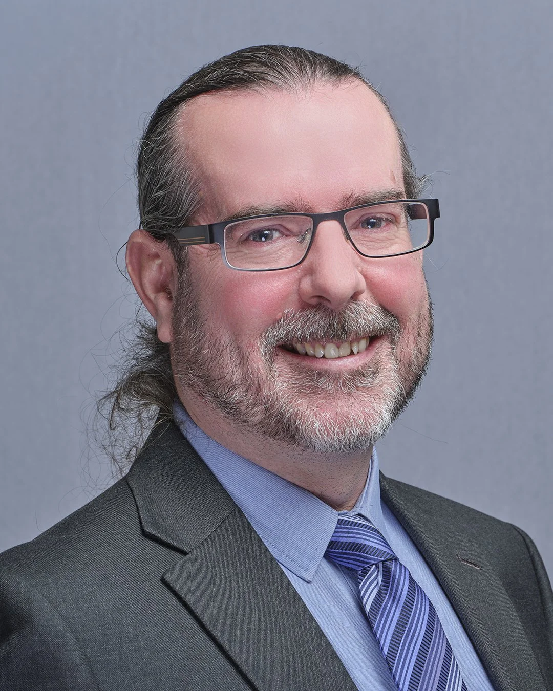 A man with gray hair, a beard, and glasses smiling, wearing a suit and tie against a gray background.