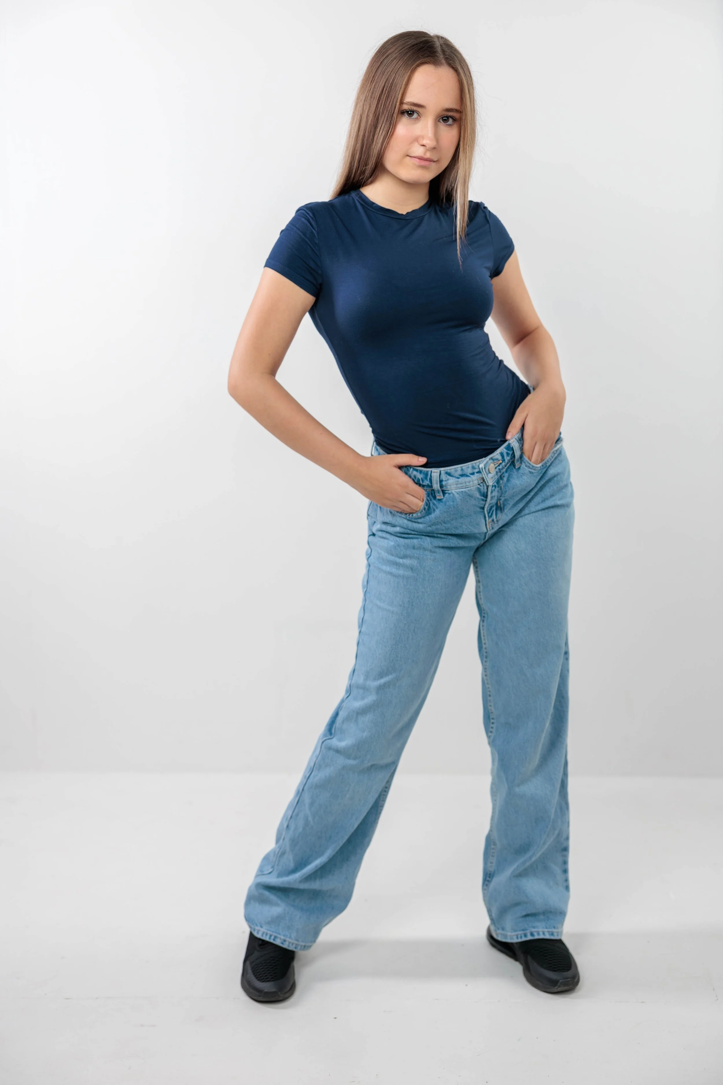 A young woman with light skin and straight, long brown hair wearing a fitted navy blue T-shirt and loose light blue jeans, standing against a plain white background with one hand in her pocket.