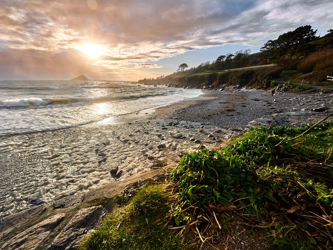 Sunset over Wembury Beach and the Mewstone in South Devon. Dramatic coastal scenery with golden sunlight hitting the waves, rocky shoreline, and green hills.