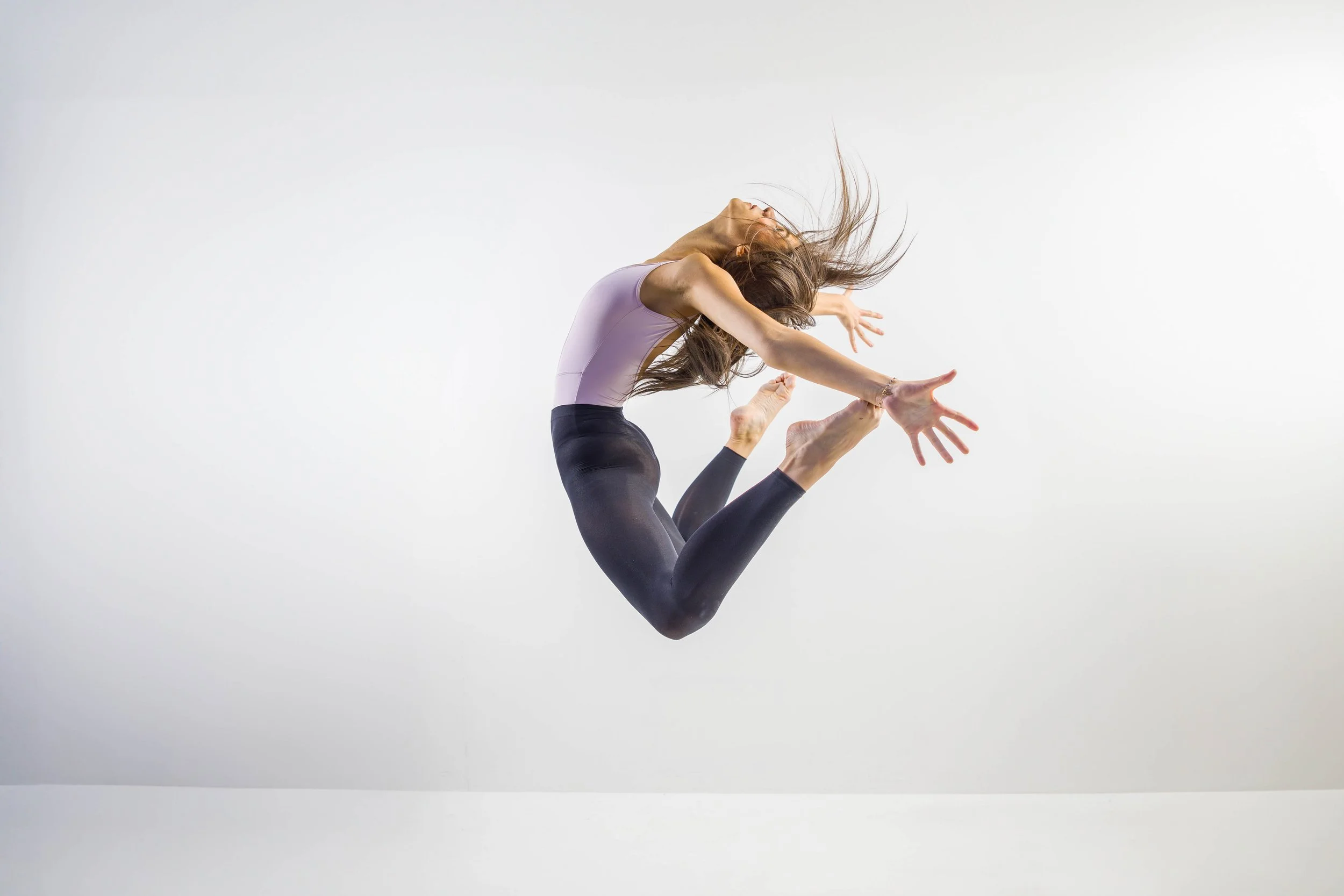 A woman in athletic wear is captured mid-air performing a dance or gymnastics move against a plain white background, with her hair flowing back and arms extended.