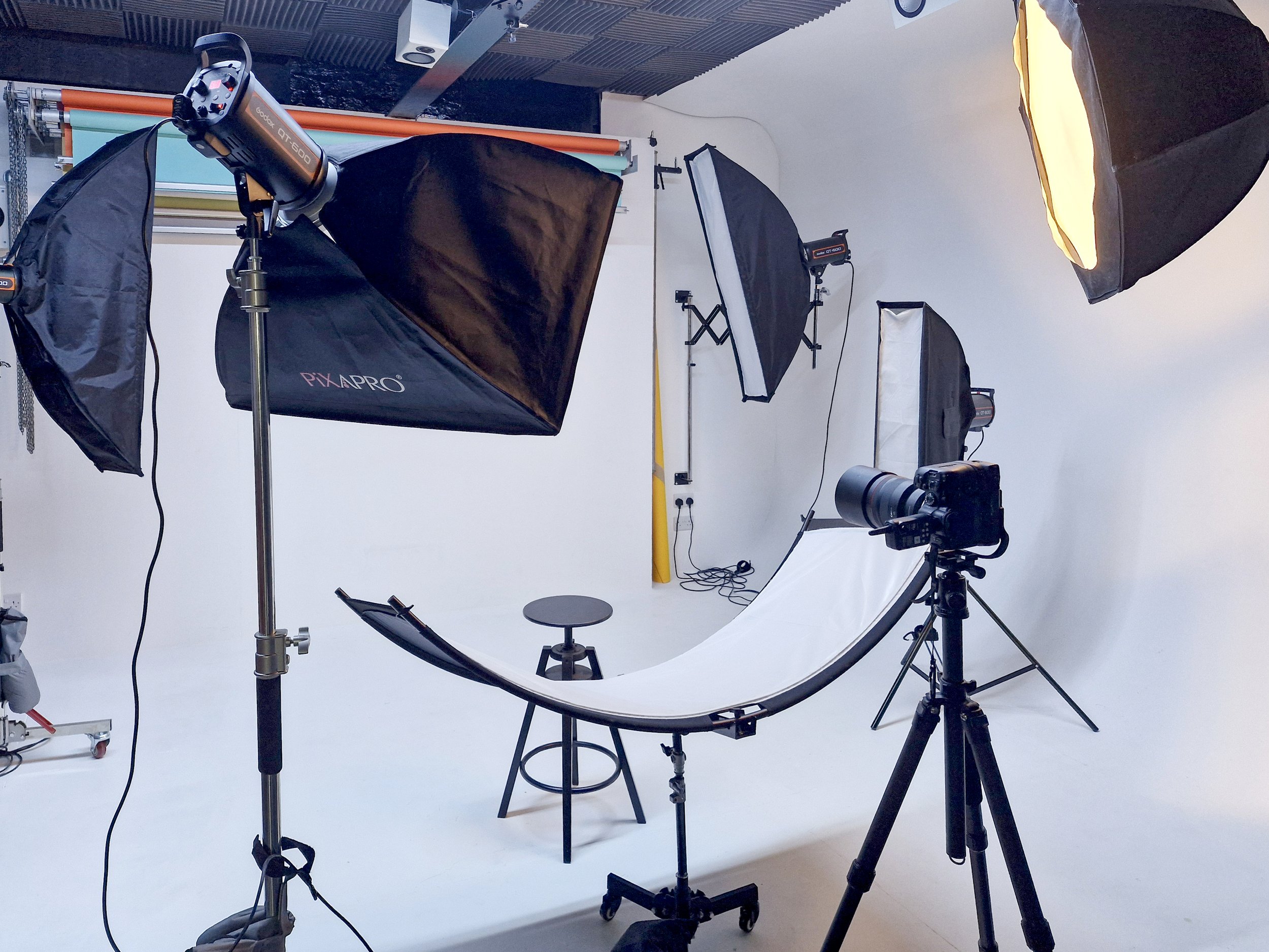 Photography studio with multiple softbox lights, a camera on a tripod, a white curved backdrop, and some chairs.