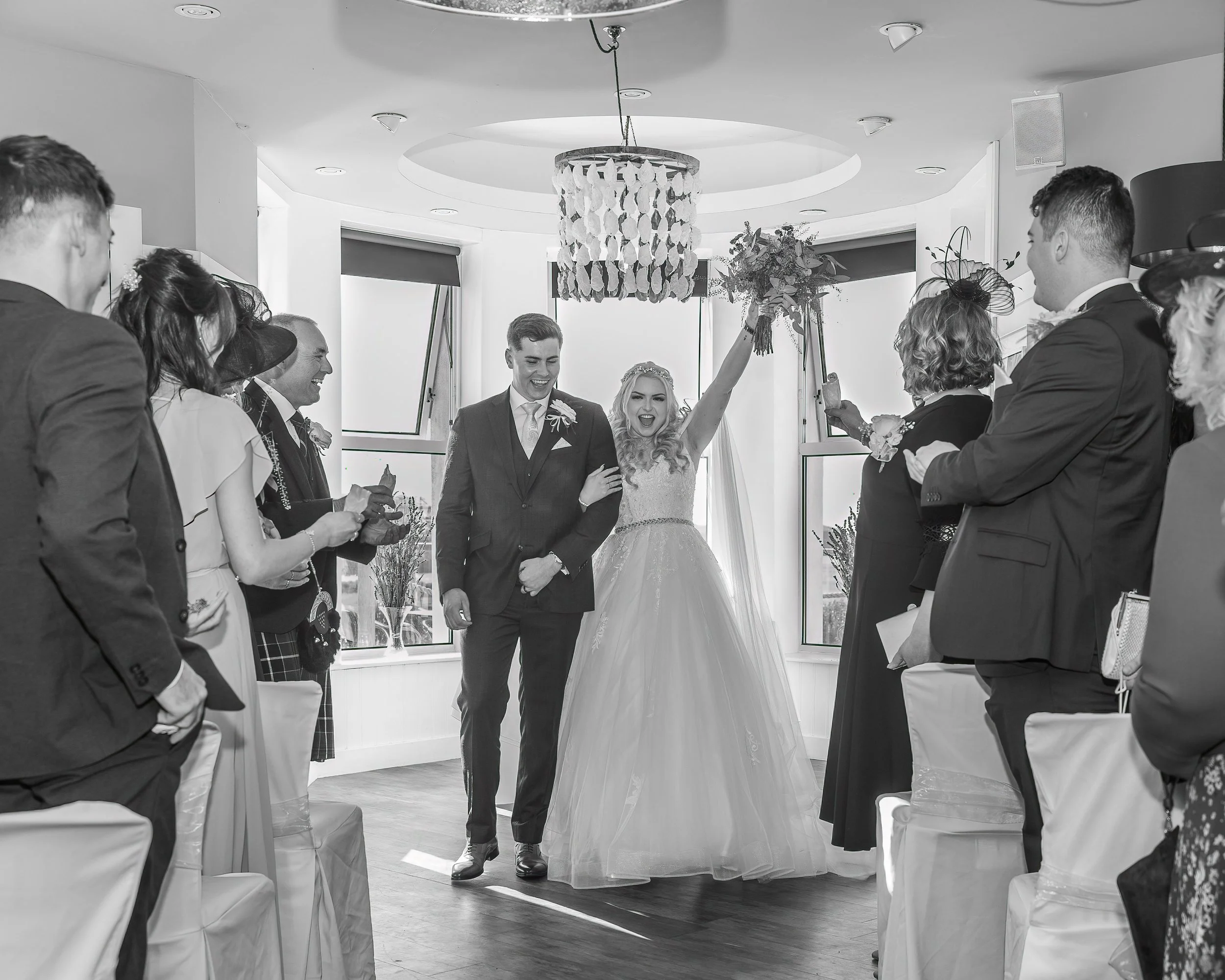 A newlywed couple joyfully enters a reception area, with the bride raising her bouquet and guests applauding. The scene is in black and white, capturing the celebration and excitement of a wedding.