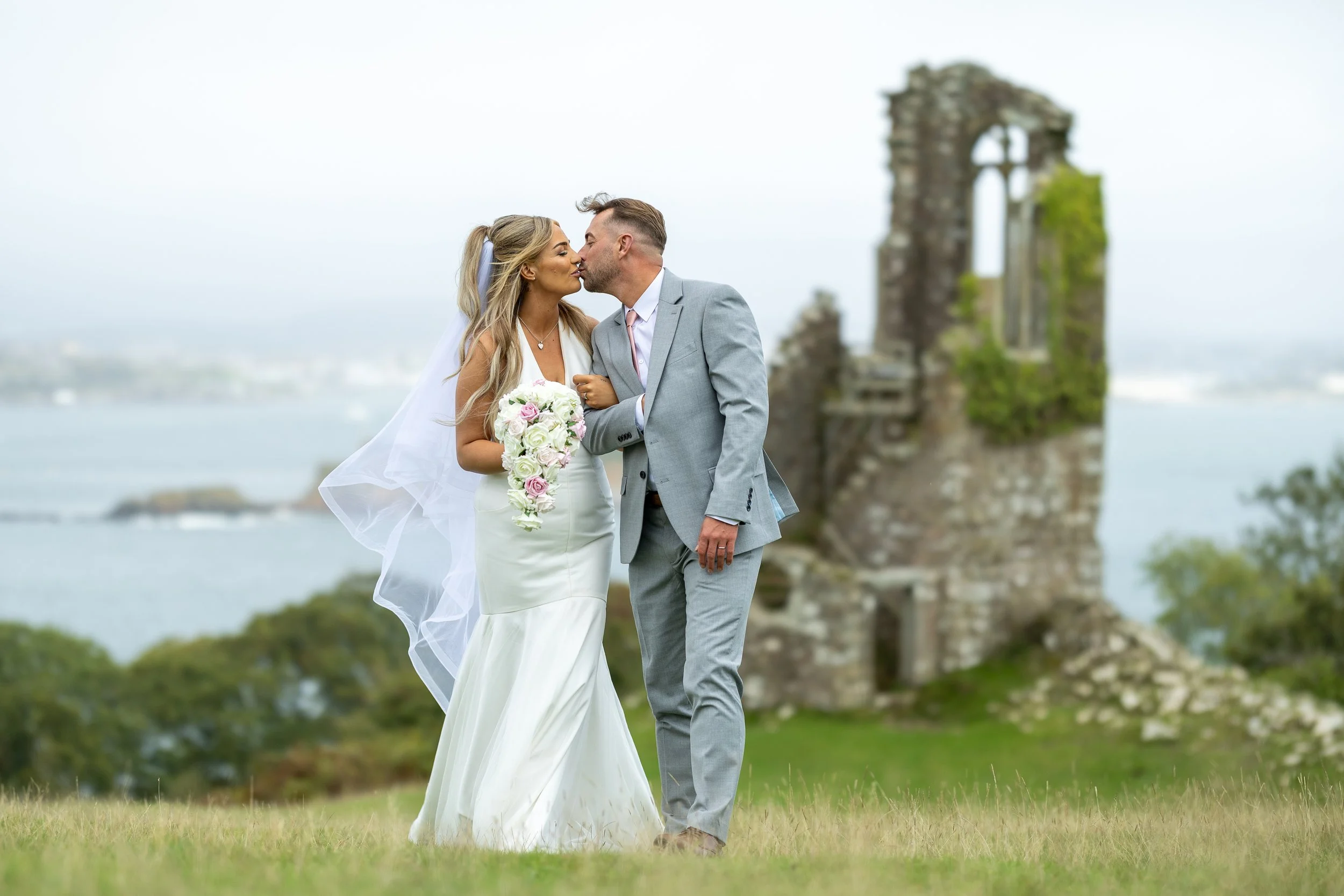 A bride and groom share a kiss outside near a ruined stone building with a body of water in the background.