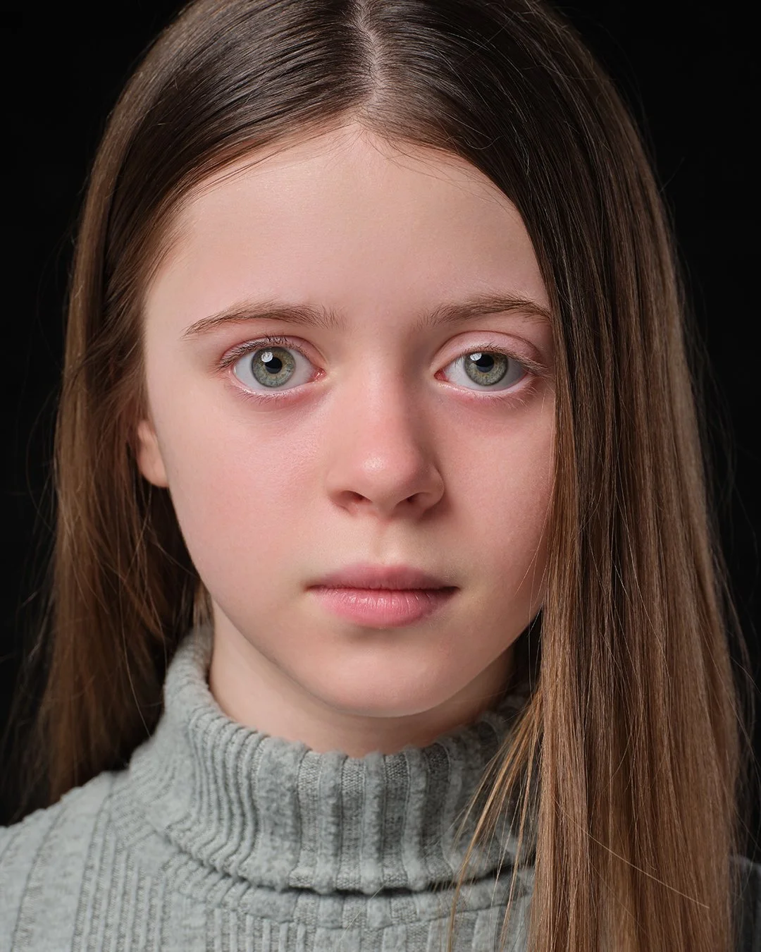 Close-up portrait of a young girl with long brown hair, fair skin, and expressive blue eyes, wearing a gray turtleneck sweater against a black background.