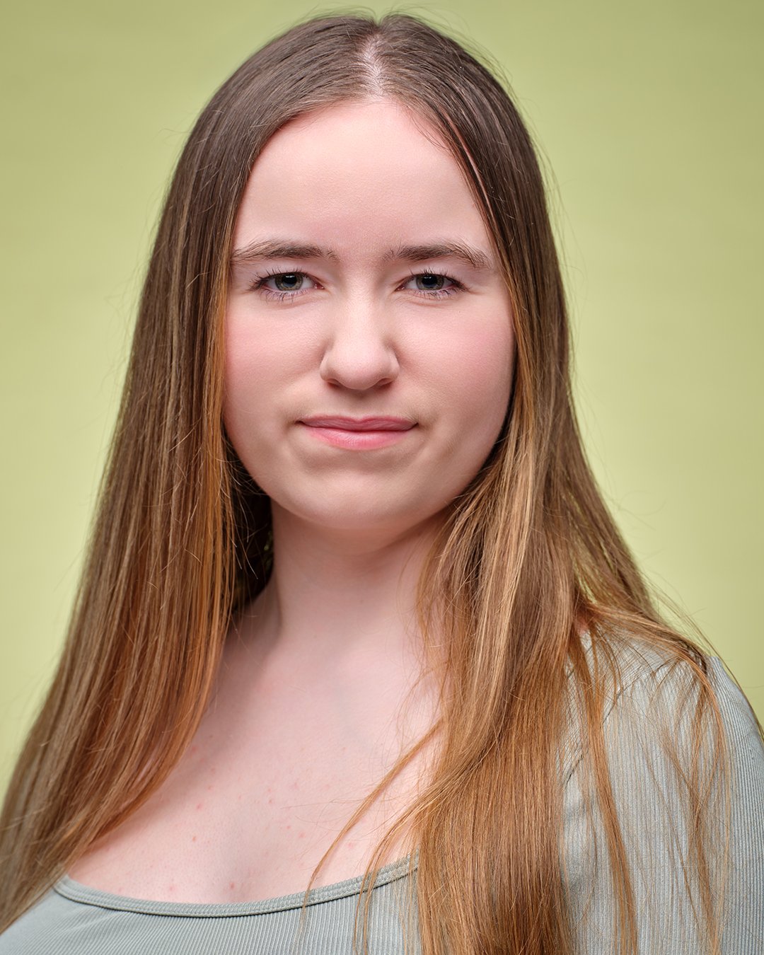 Studio headshot of a young woman with long light brown hair wearing a sage green top, captured by a professional portrait photographer against a soft green background.