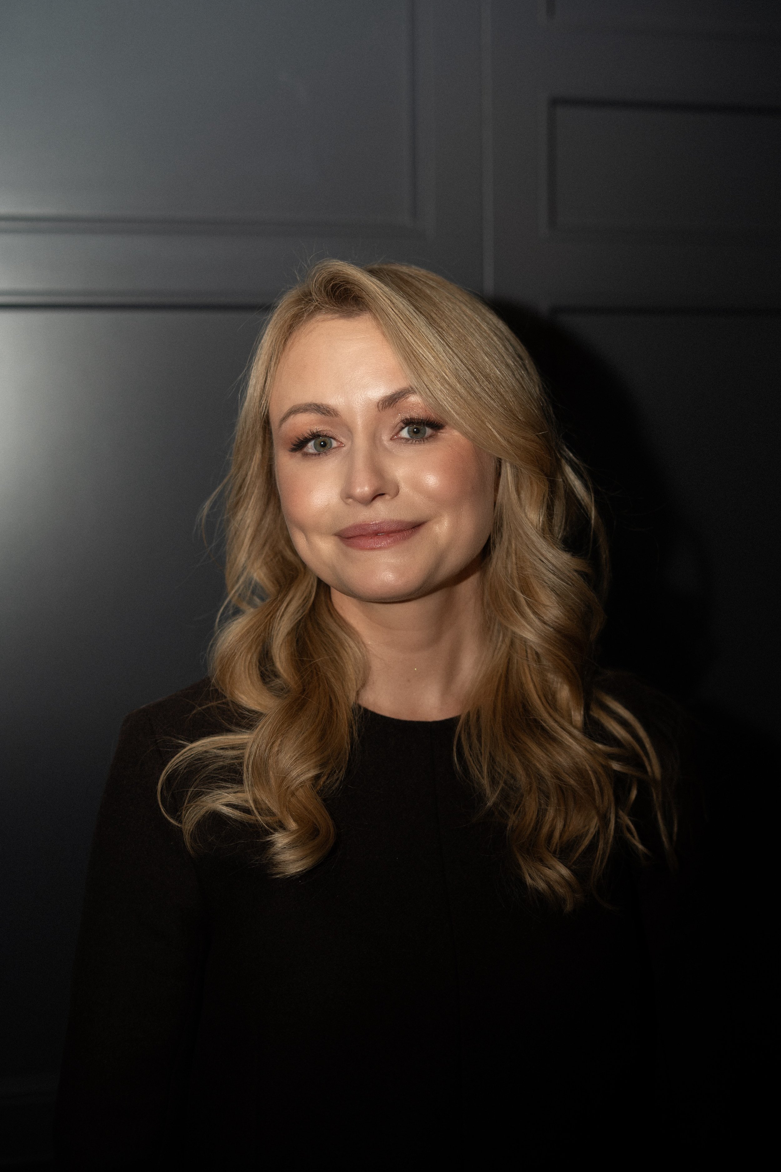 Frau im dunklen Kleid, blondem, gelocktem und schulterlangem Haar vor einer dunklen Wand.