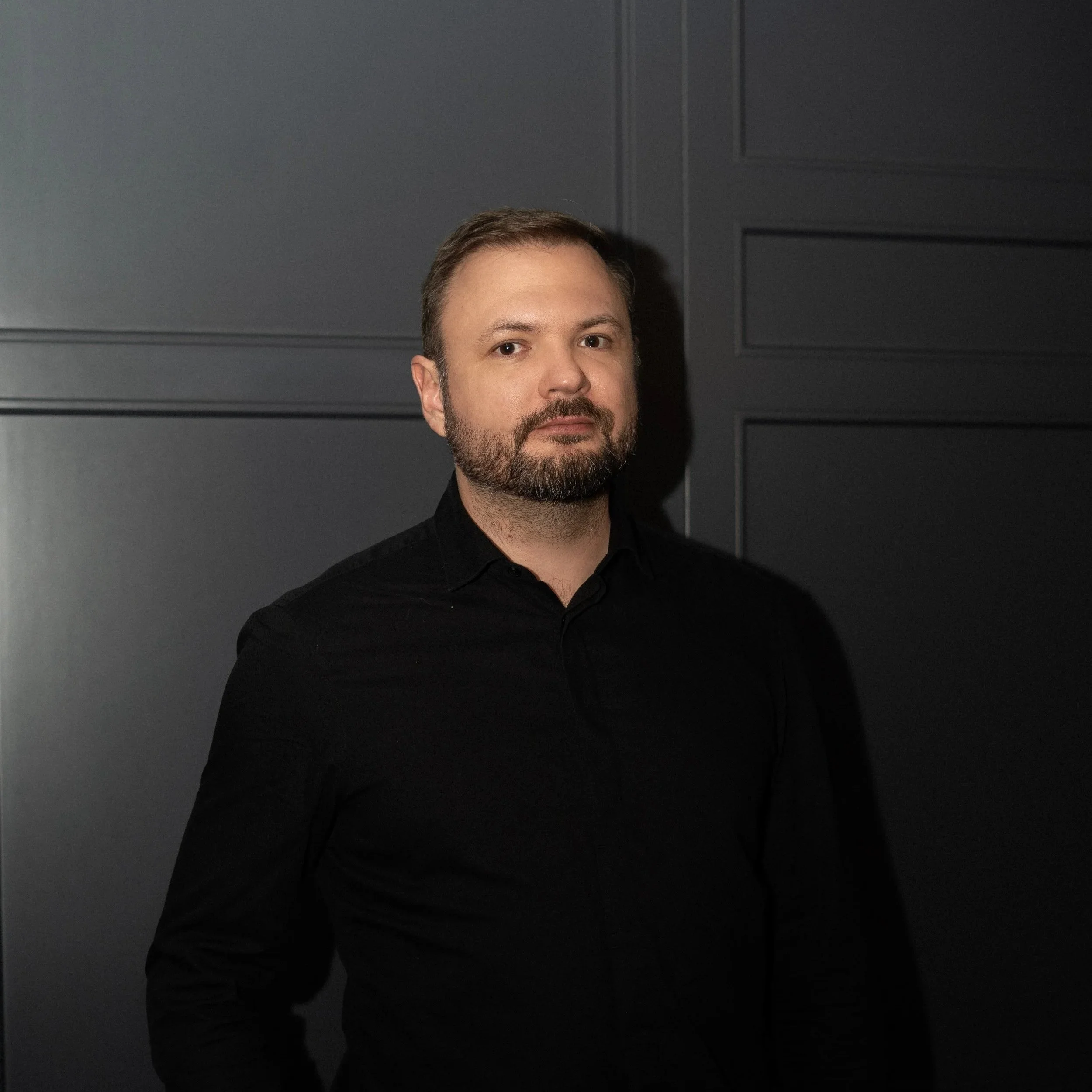 Mann mit Schnurrbart und kurzem Bart im schwarzen Hemd vor einer dunklen Wand.